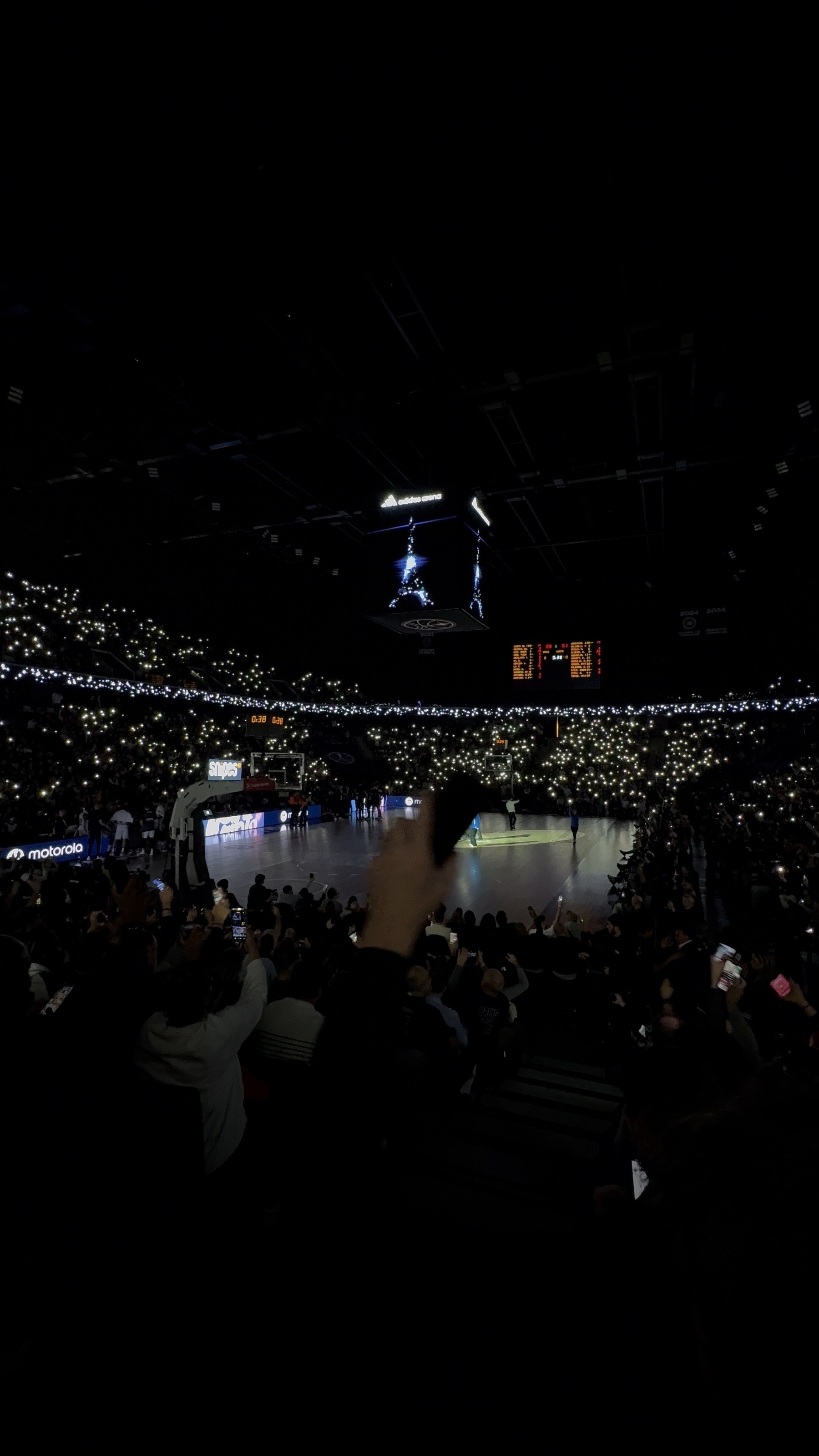 A darkened stadium is filled with spectators holding up lit devices, creating a starry effect as they watch a basketball game on the illuminated court.