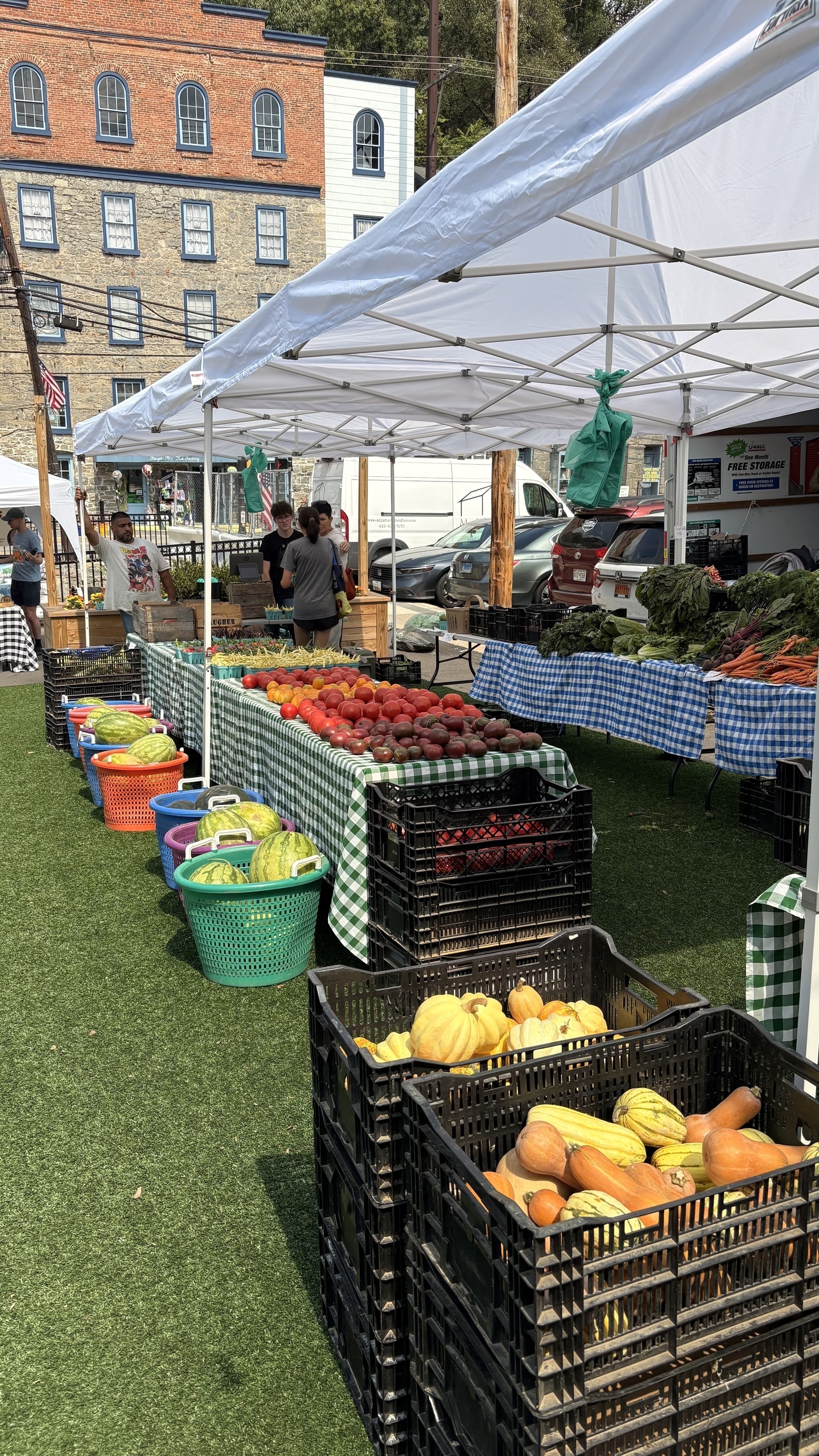 A farmer's market stall displays an array of fresh vegetables and fruits under white tents, with colorful baskets and brick buildings in the background.