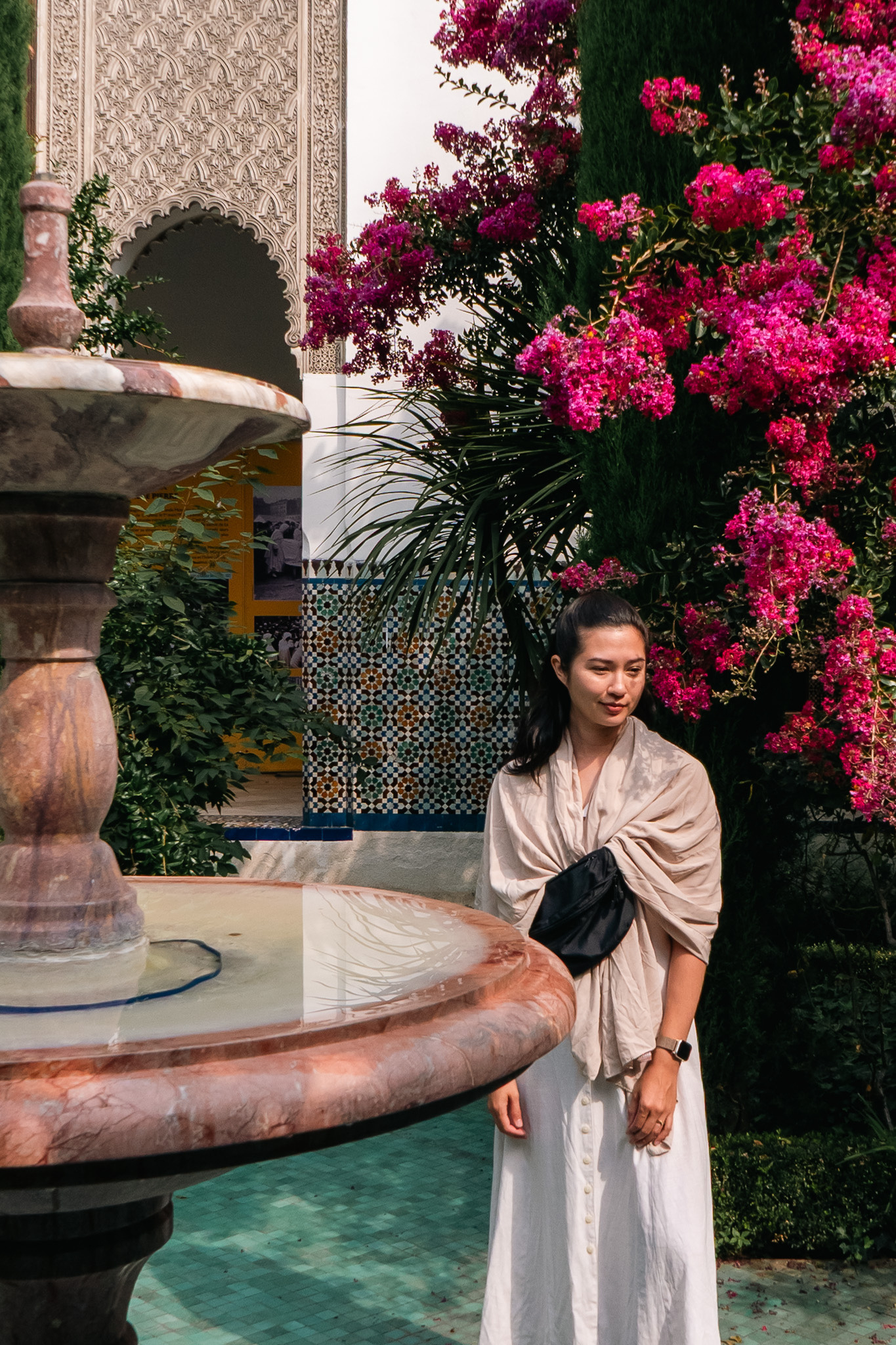 A woman stands near a decorative fountain surrounded by vibrant pink flowers and intricate architecture.