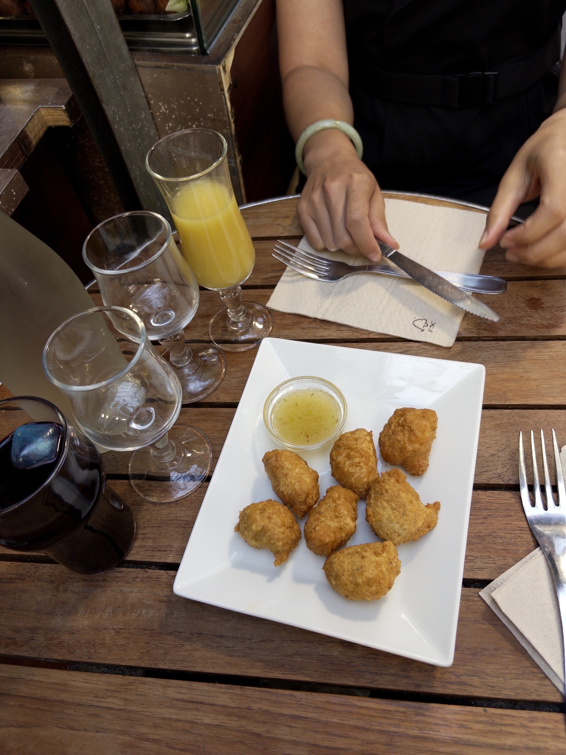 A plate of fried appetizers with dipping sauce is set on a wooden table alongside glasses and someone preparing to eat with a knife and fork.