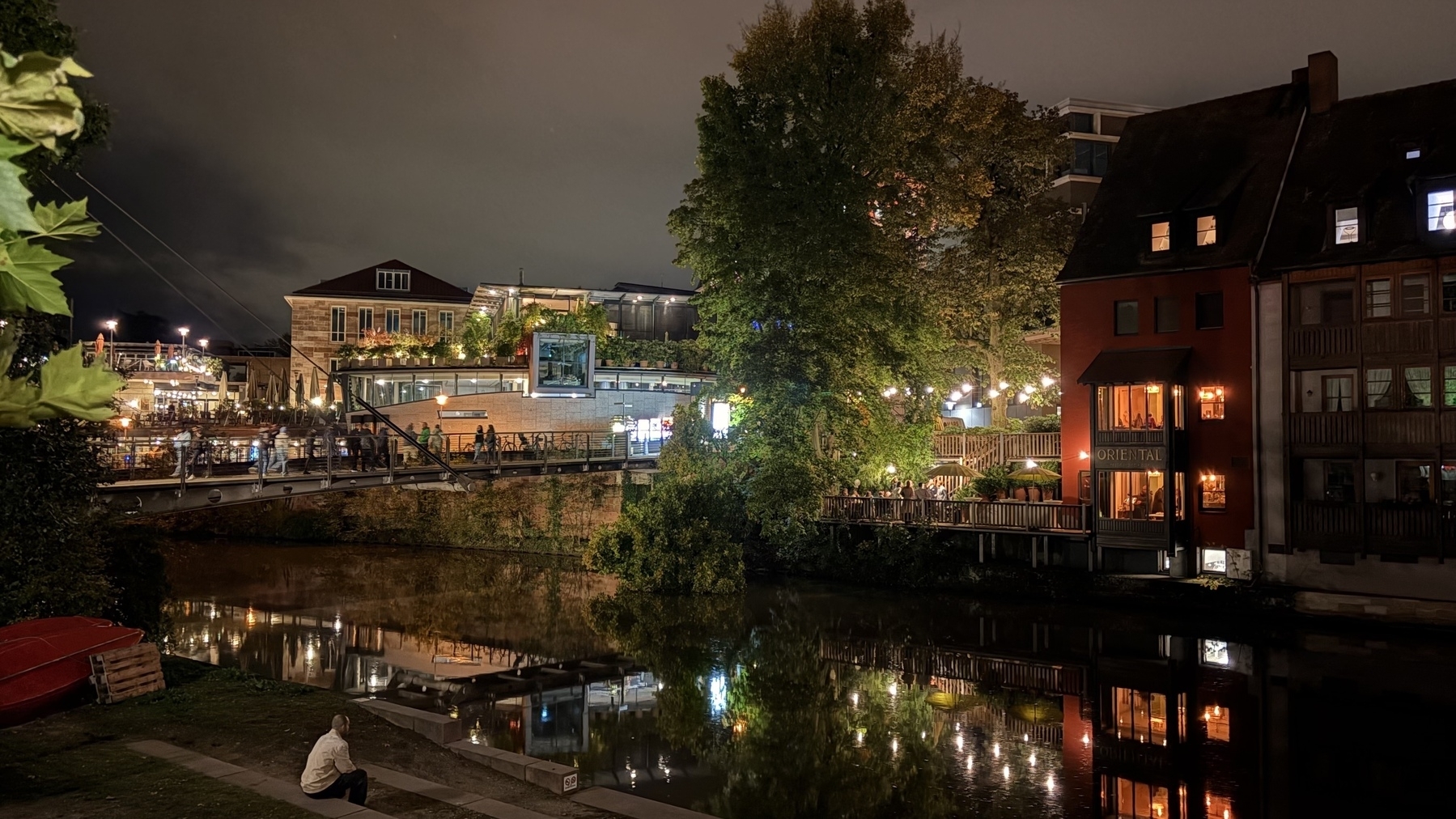 A peaceful night scene by a river features illuminated buildings, a bridge with people, and a person sitting near the water.