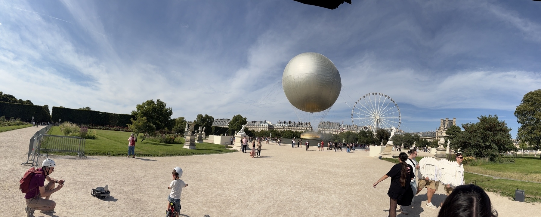 A park scene shows people walking and gathering near a large reflective sphere and a Ferris wheel under a partly cloudy sky.