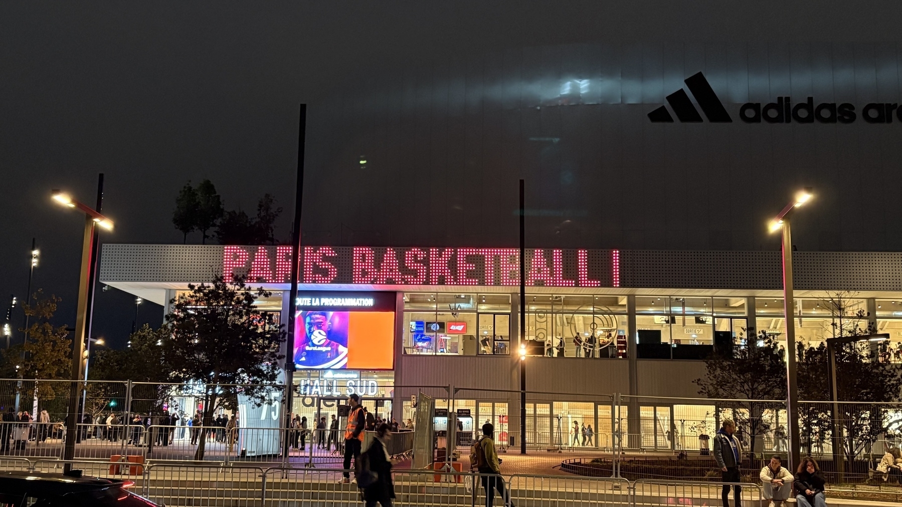 A brightly lit building features the text PARIS BASKETBALL with people walking nearby and a large screen displaying an advertisement.