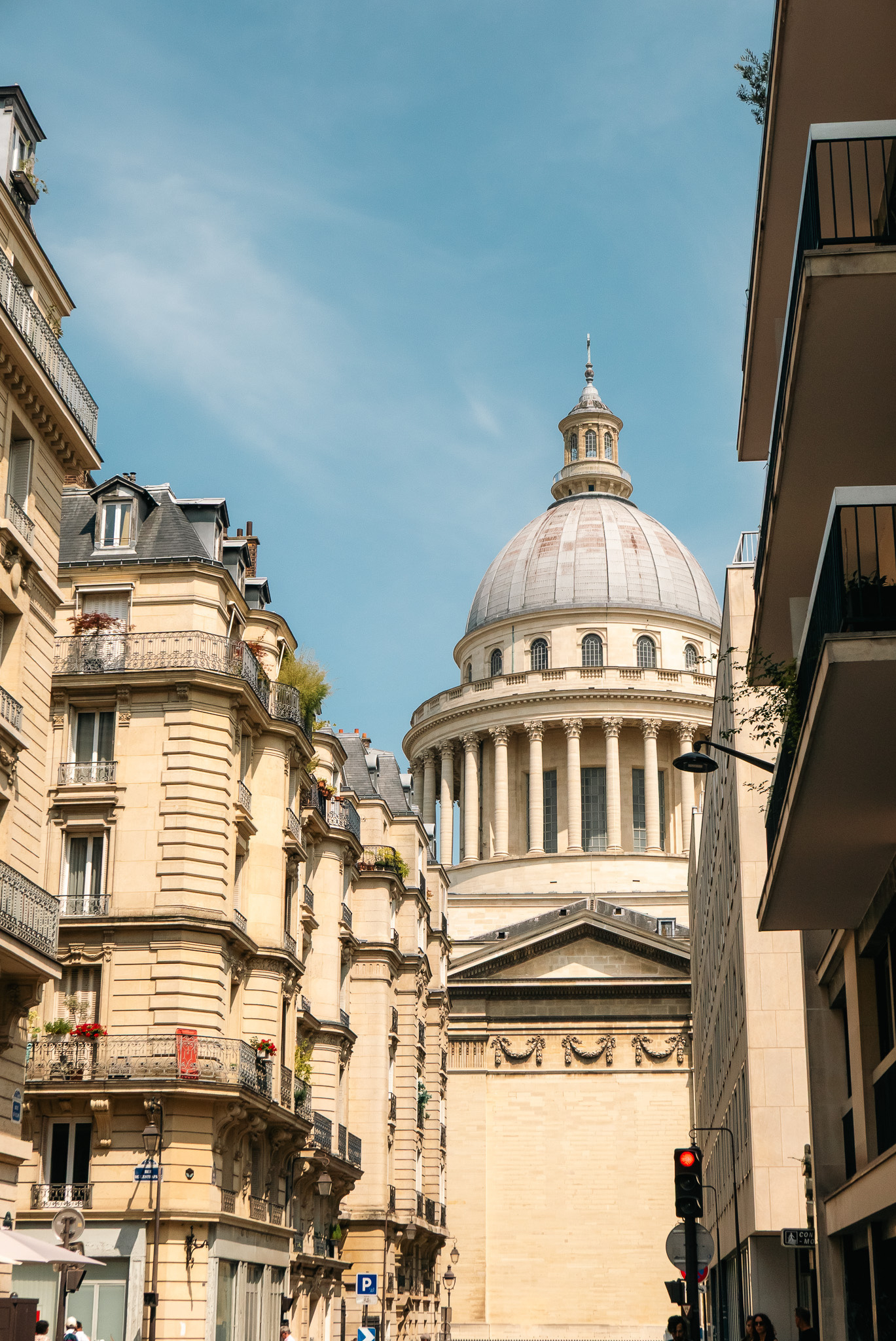 A street with classic European architecture leads to a grand domed building under a clear blue sky.