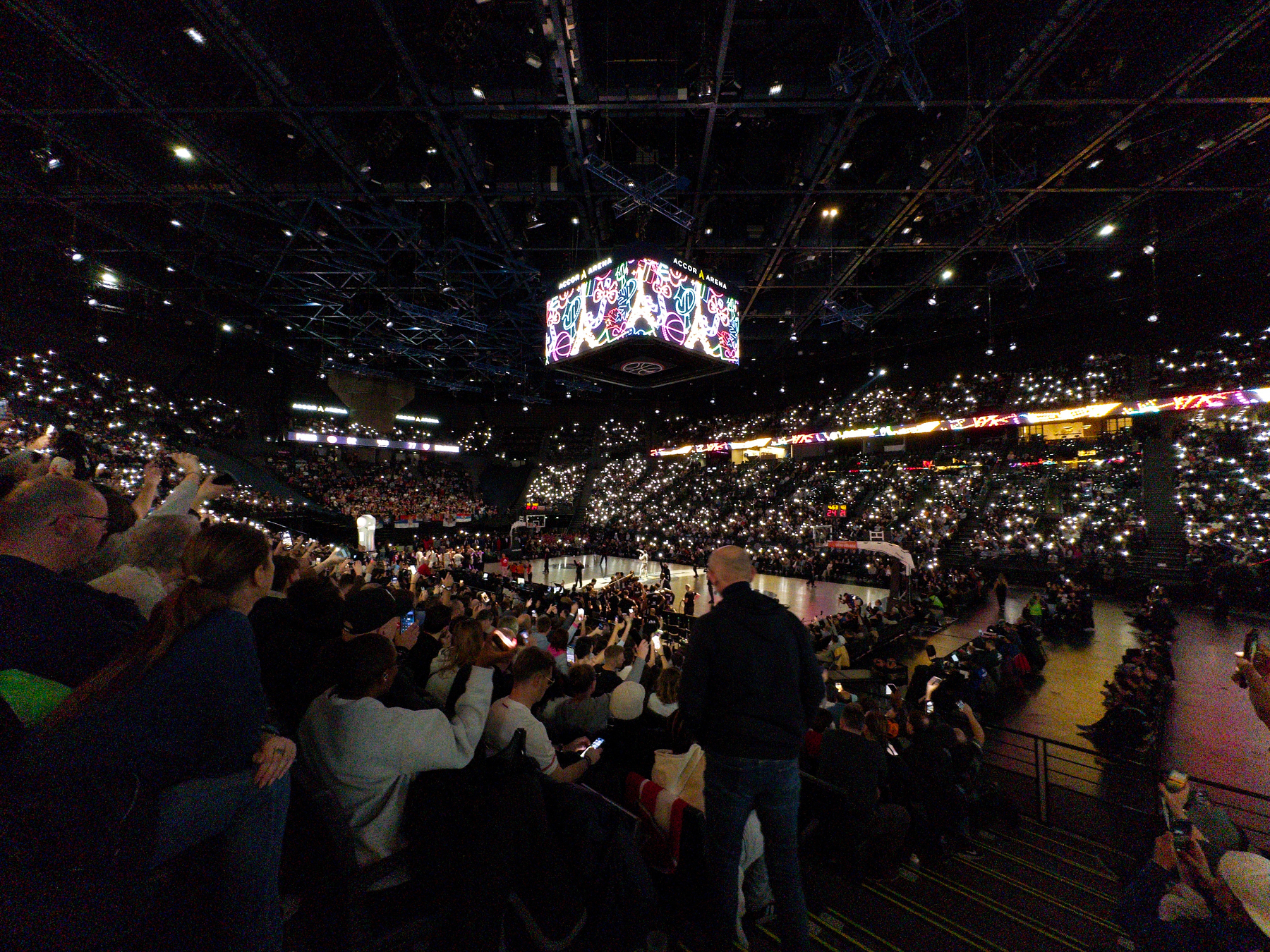 A large crowd in an arena is illuminated by the glow of countless phone screens, with a central display hanging above the event.