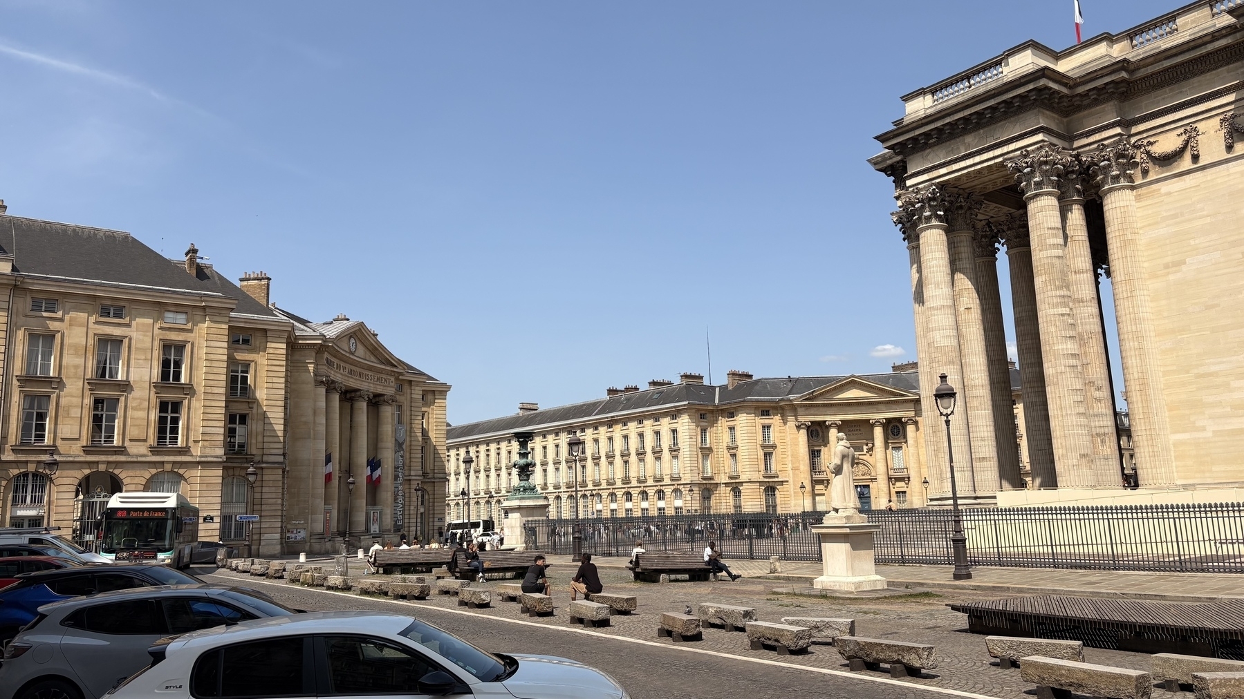 A grand architectural scene features historic buildings with tall columns under a clear blue sky, alongside parked cars and a few people strolling nearby.