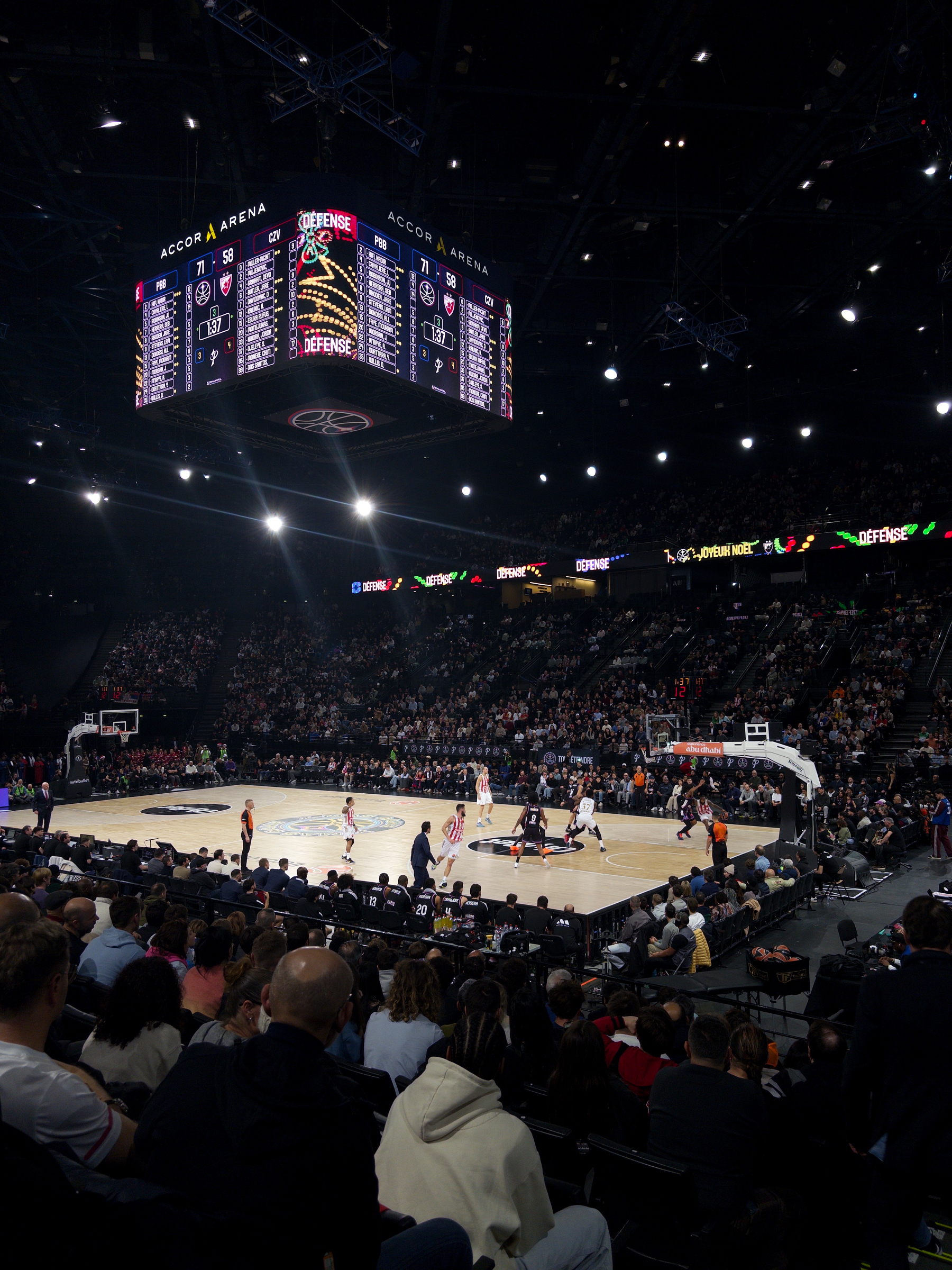 A large crowd watches a basketball game in progress at Accor Arena, with a scoreboard hanging overhead.