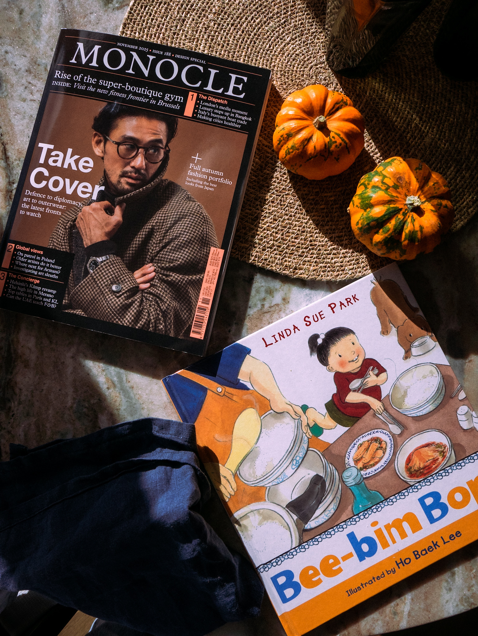 A Monocle magazine, a children's book titled Bee-bim Bop! by Linda Sue Park, small pumpkins, and a cloth are arranged on a surface.