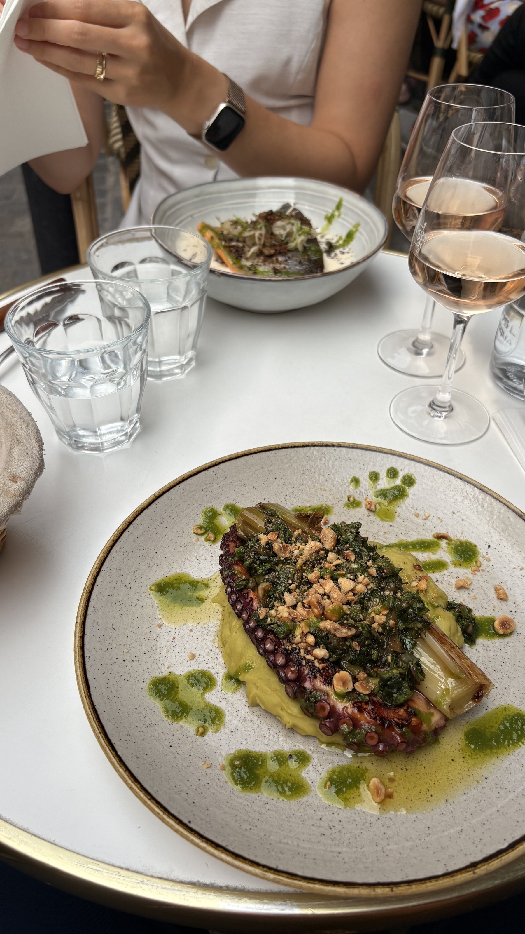 A meal featuring a dish with octopus and garnishes is served on a white table alongside glasses of water and wine, with a person in the background.