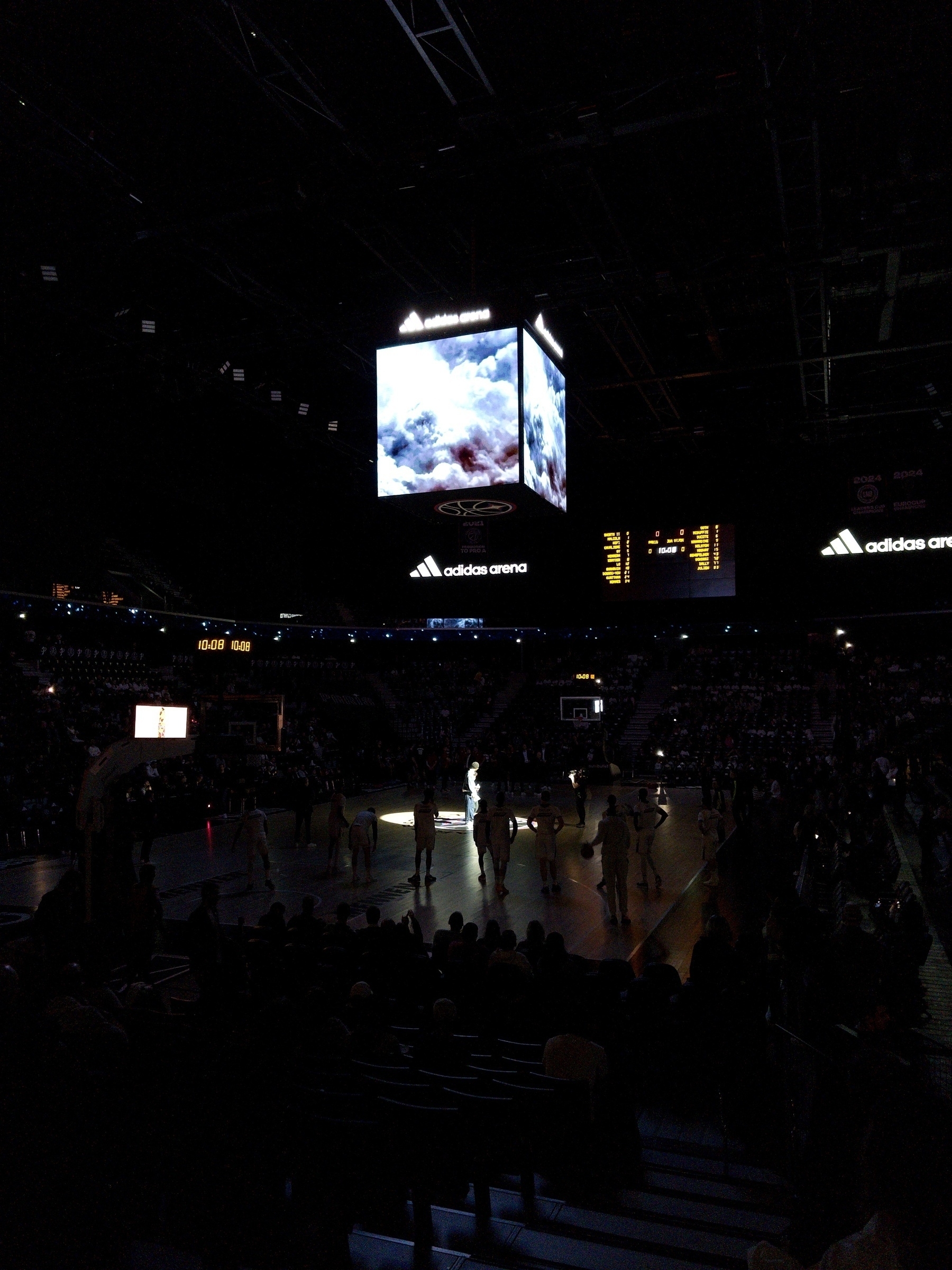 A basketball game is taking place in an arena with a spotlight on the players and a large electronic screen overhead.