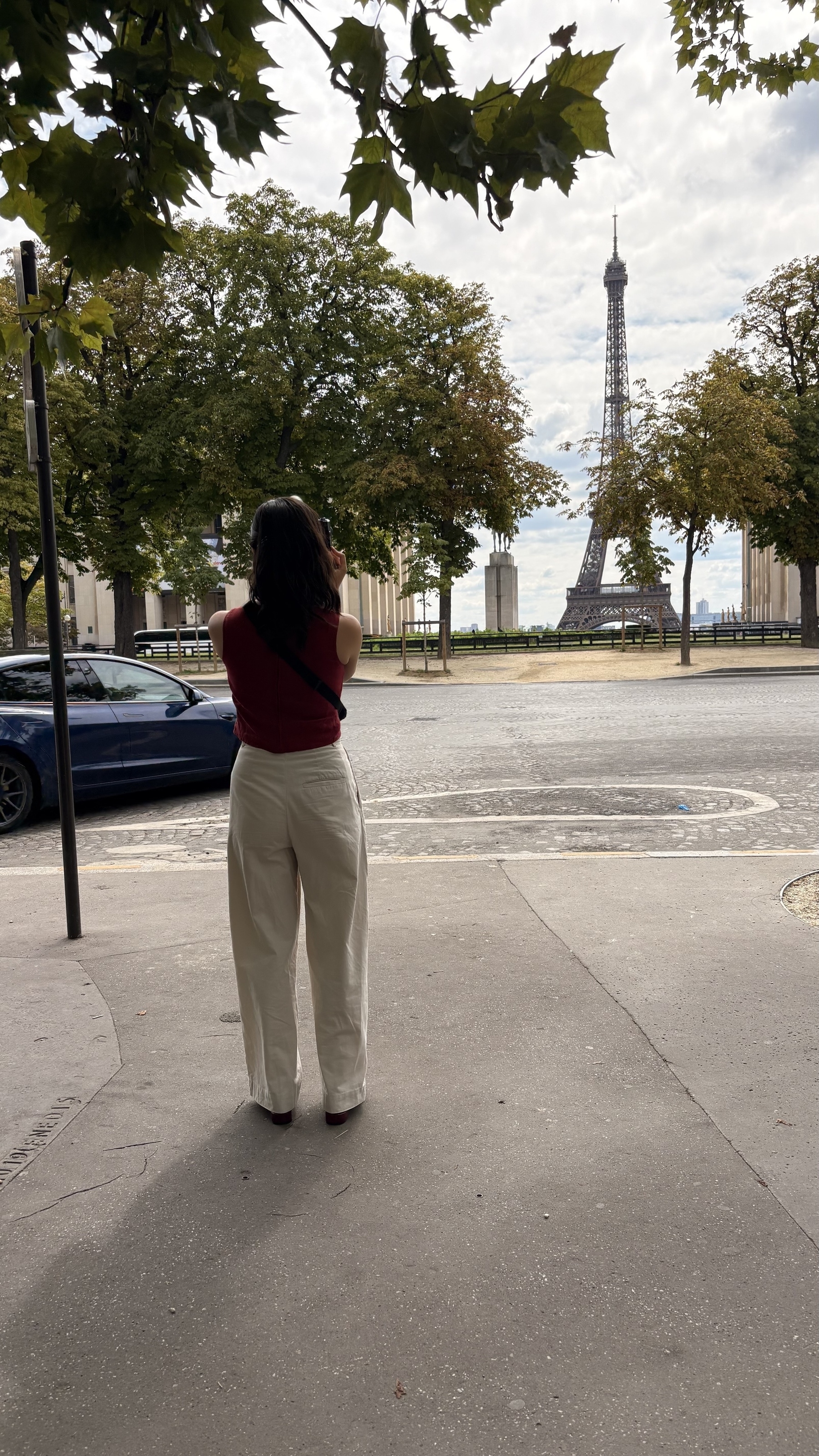 A person is standing on a sidewalk taking a photo of the Eiffel Tower in the distance, surrounded by trees and urban scenery.