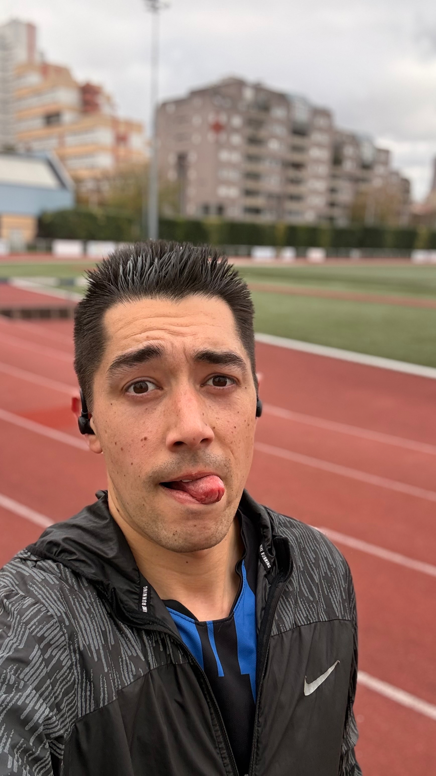 A person in athletic gear with earbuds is on a running track with buildings in the background.