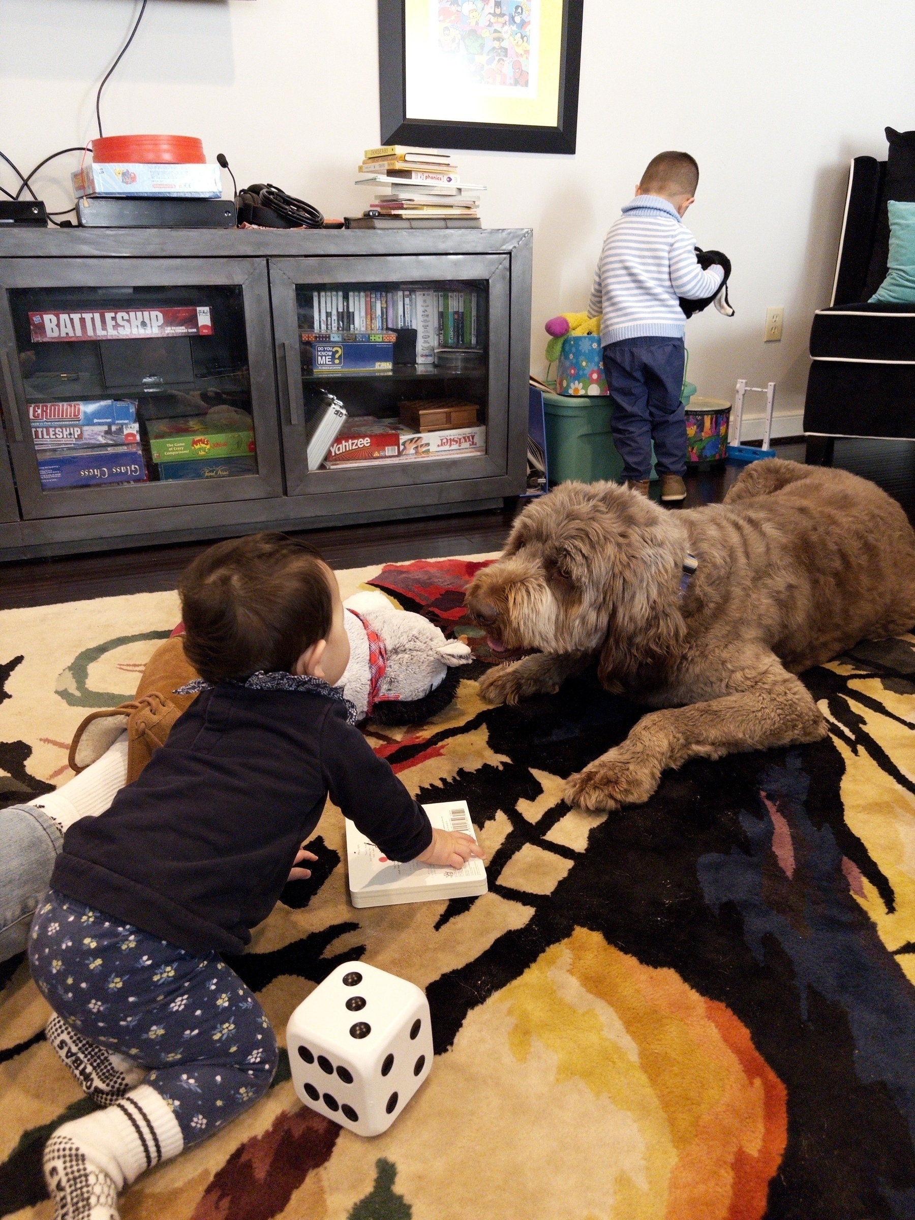 A cozy living room scene shows two children playing with toys on a colorful rug alongside a fluffy dog, with board games and books visible on a nearby shelf.