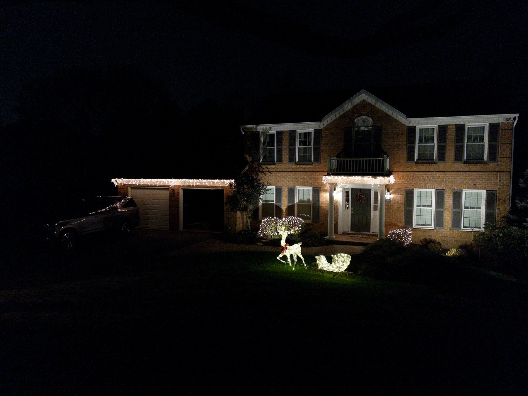 A brick house is adorned with festive white lights and features illuminated reindeer decorations on the front lawn at night.