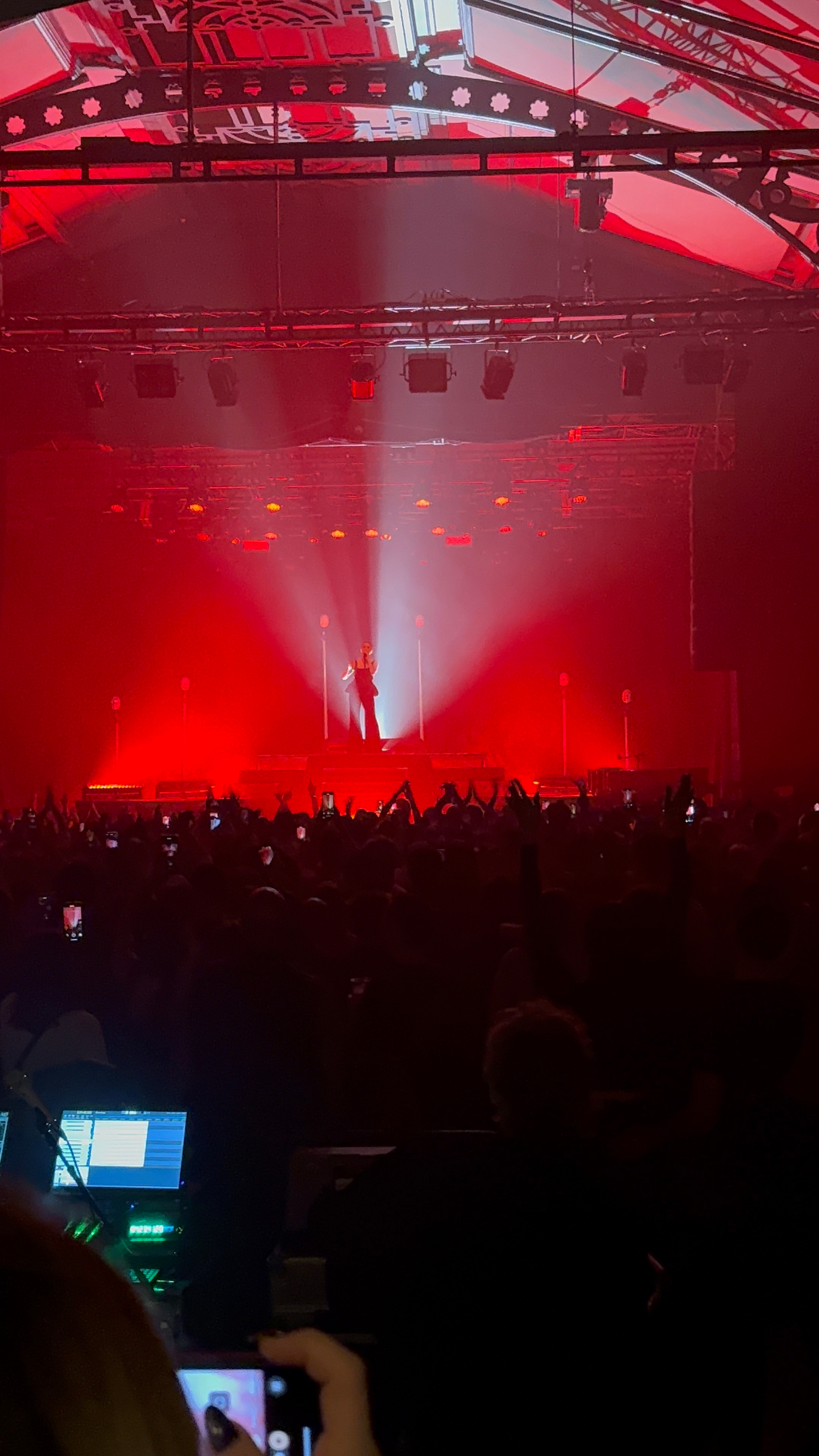 A performer stands on stage under dramatic red lighting in front of a large crowd at a concert venue.