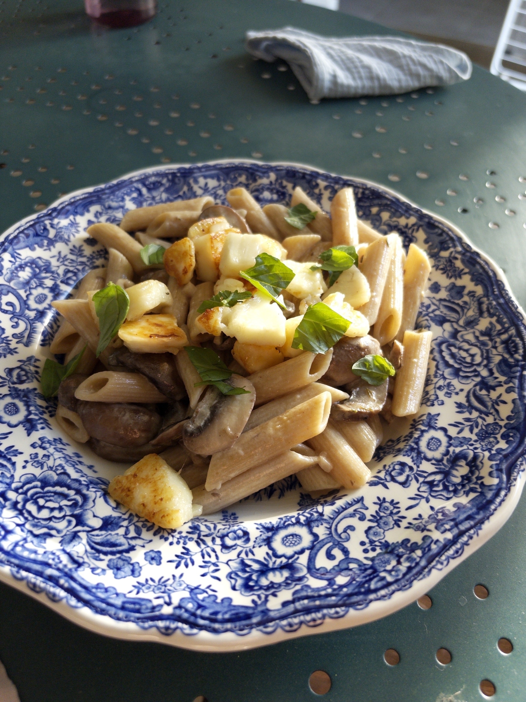 A plate of penne pasta is topped with mushrooms, cheese, and fresh basil, served on a decorative blue and white dish.