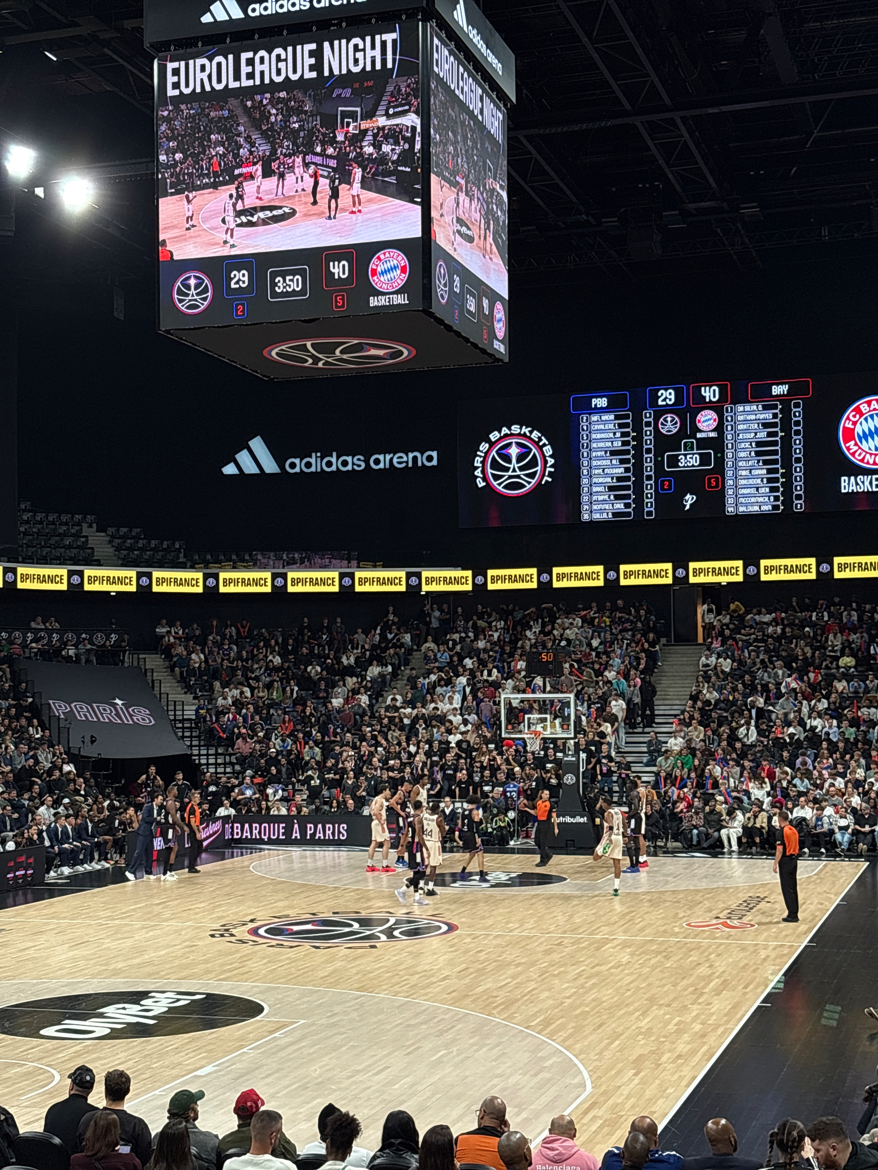 A Euroleague basketball game is taking place in a packed arena with a large overhead scoreboard displaying the score and advertisements.