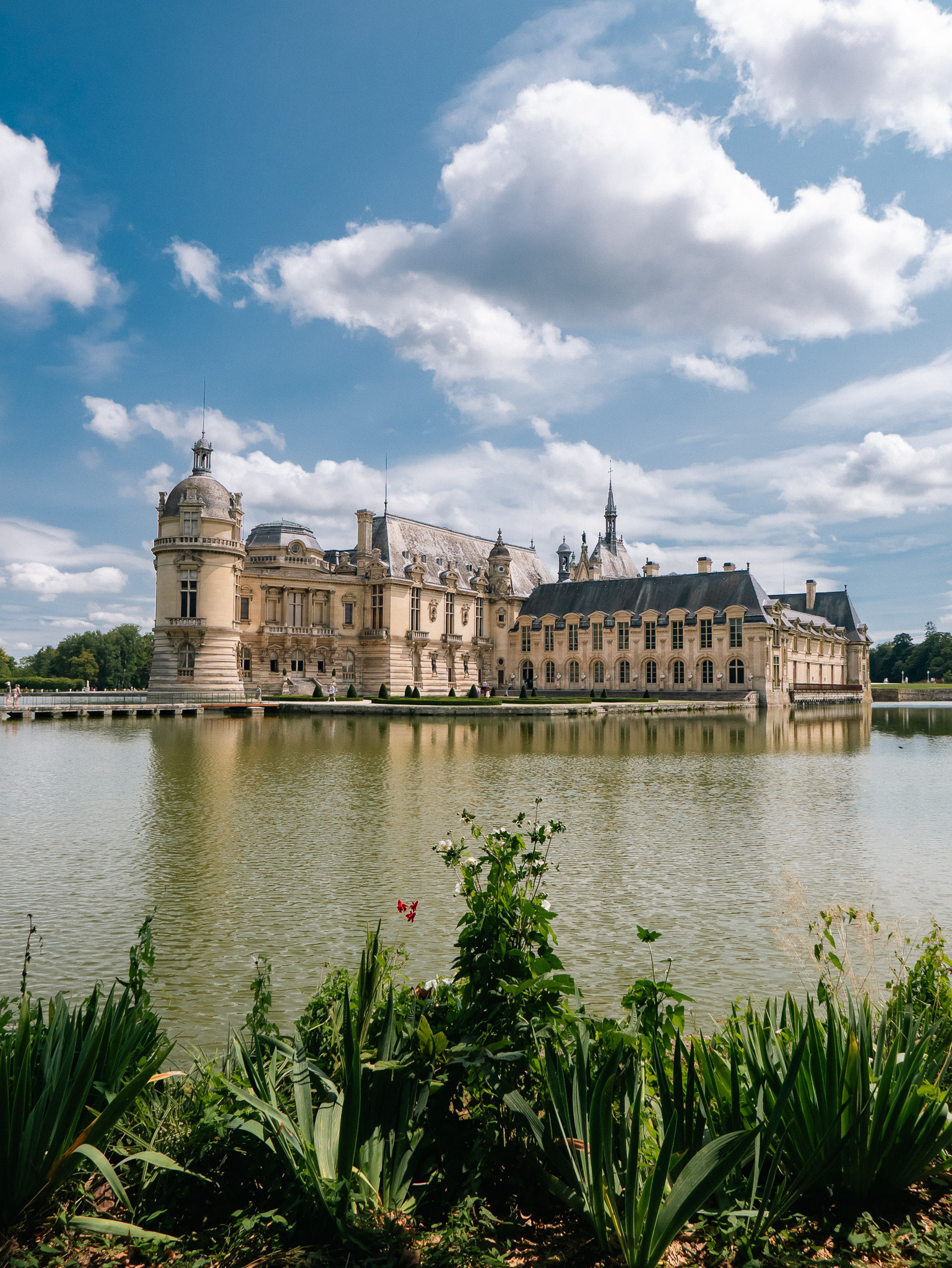 A majestic castle stands beside a serene lake under a partly cloudy sky, surrounded by lush greenery.