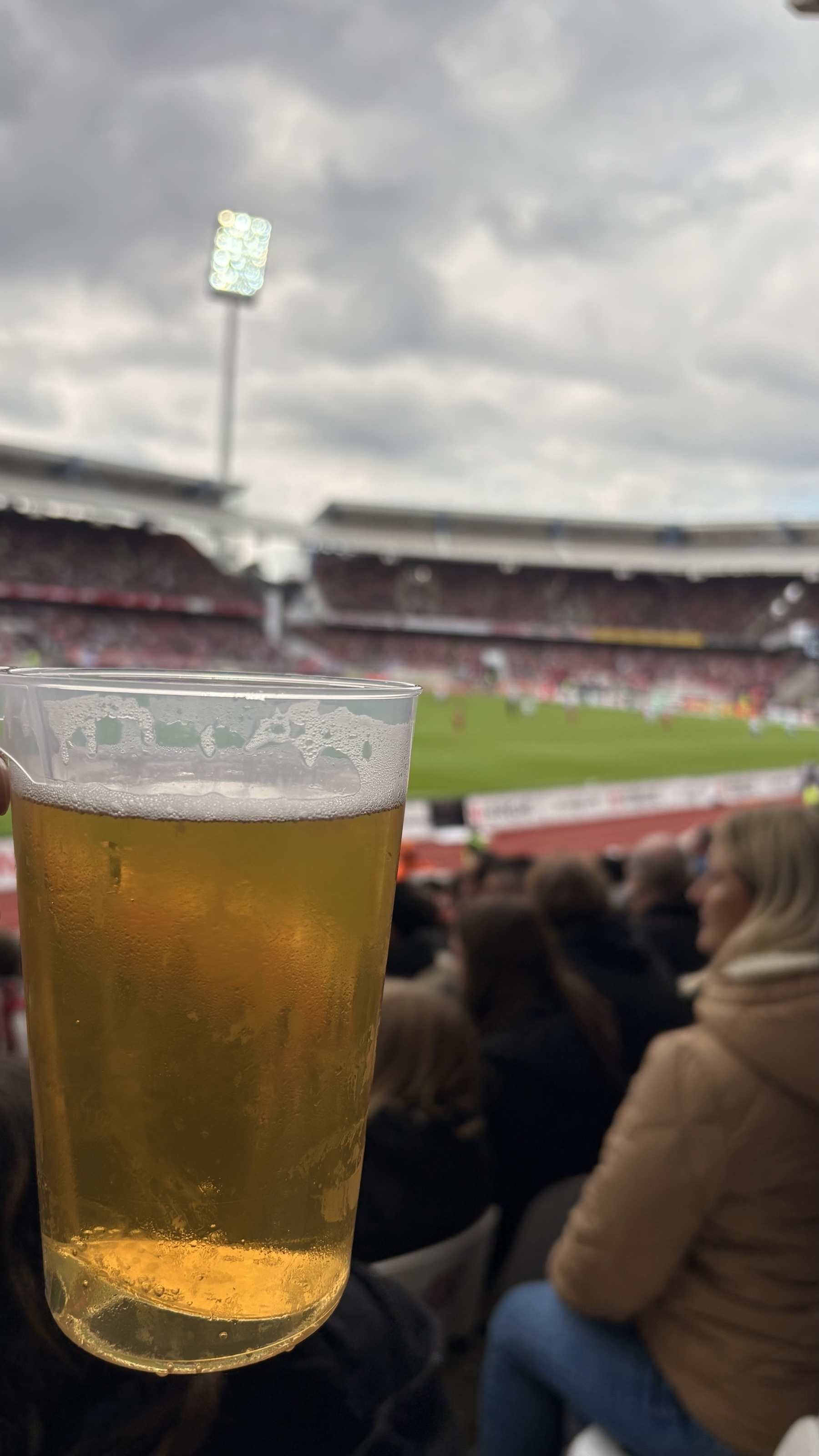 A person is holding a plastic cup of beer at a crowded sports stadium with a soccer match in progress.
