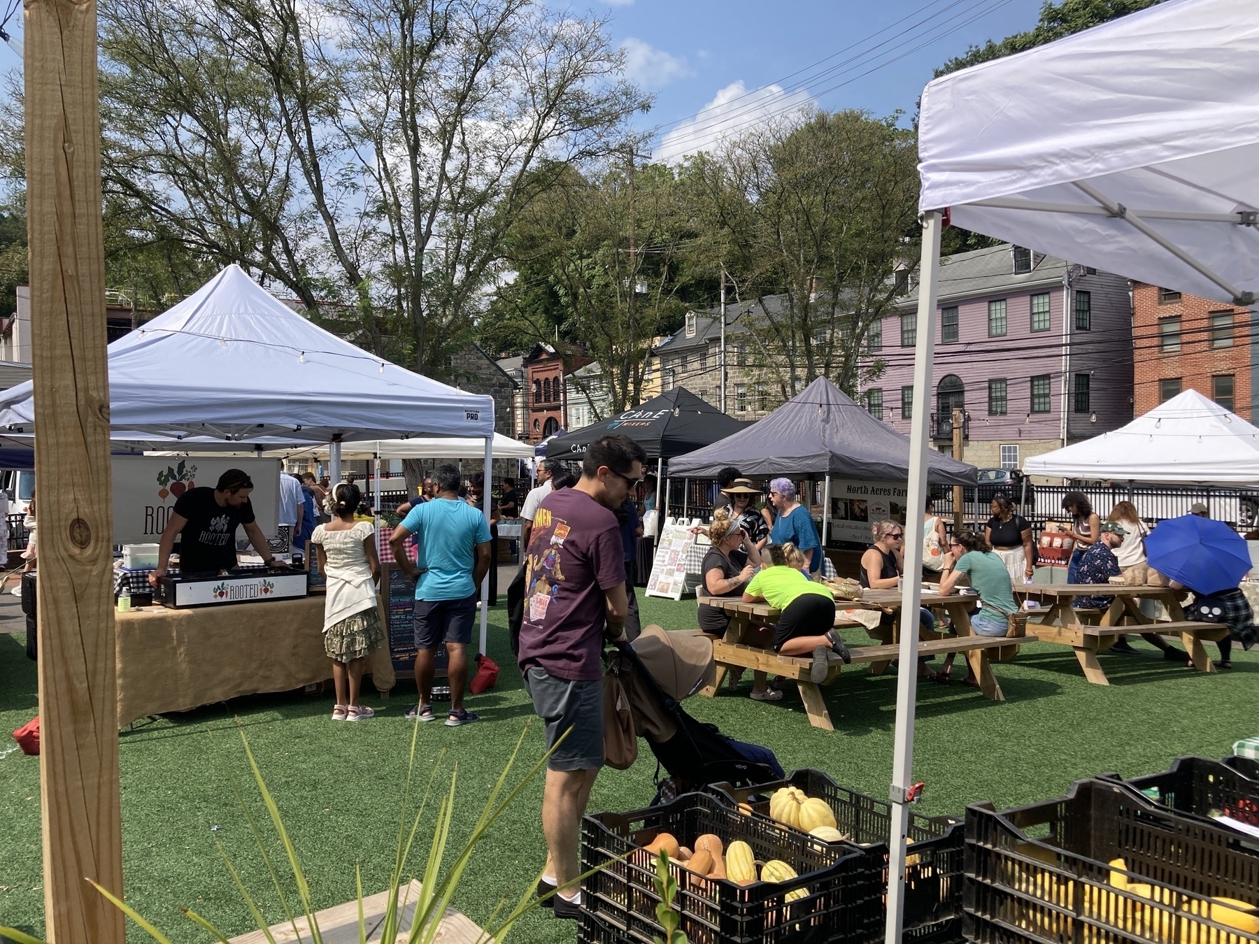 People are enjoying a sunny day at an outdoor market with tents, food stalls, and picnic tables.