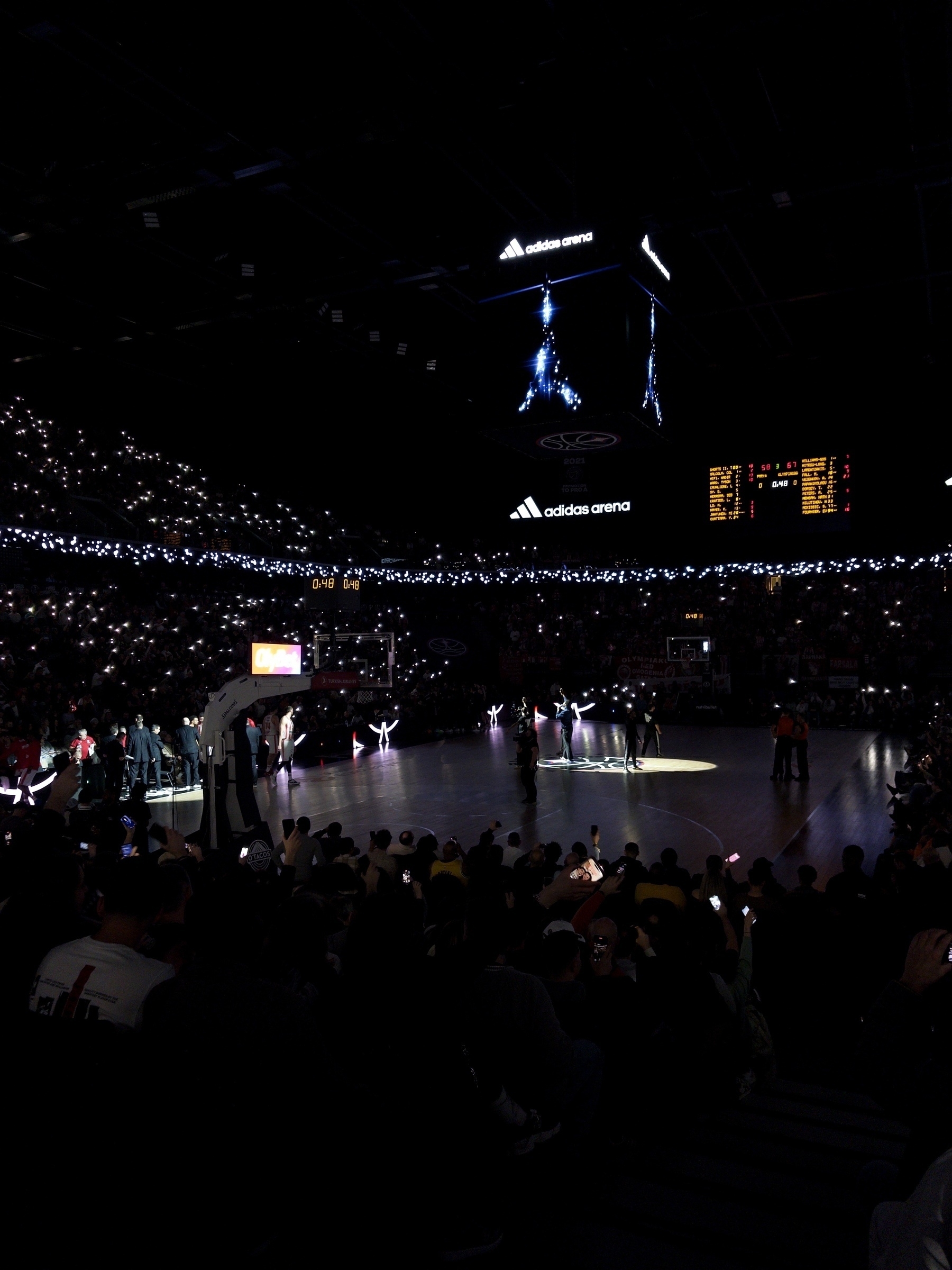 A basketball court illuminated by stage lighting and surrounded by a darkened stadium full of spectators, with performers or players spotlighted at the center.