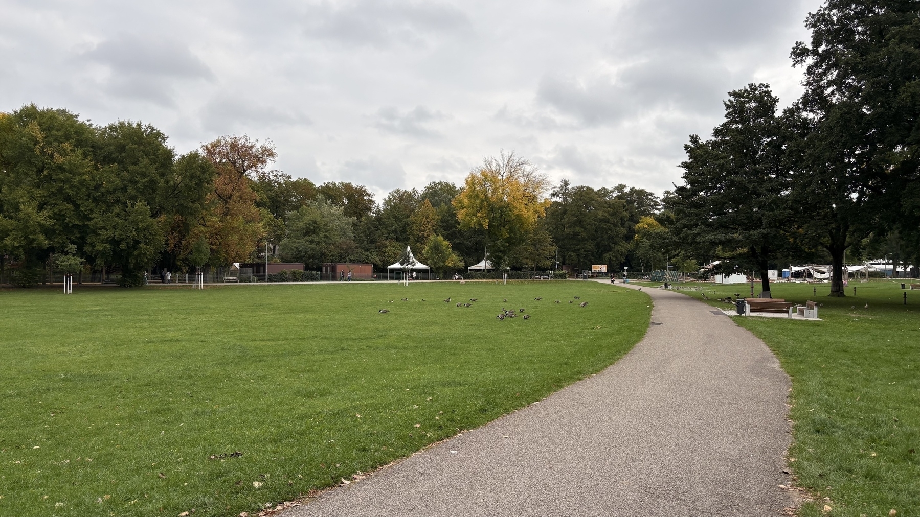 A path winds through a park with lush green grass and trees under a cloudy sky.