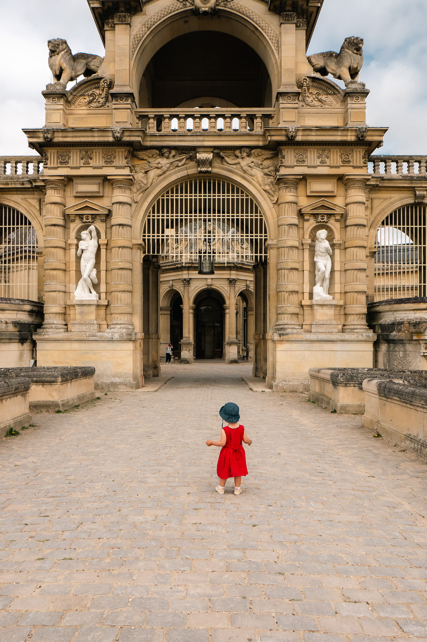 A child in a red dress walks towards an ornate, historic building with statues and architectural details.