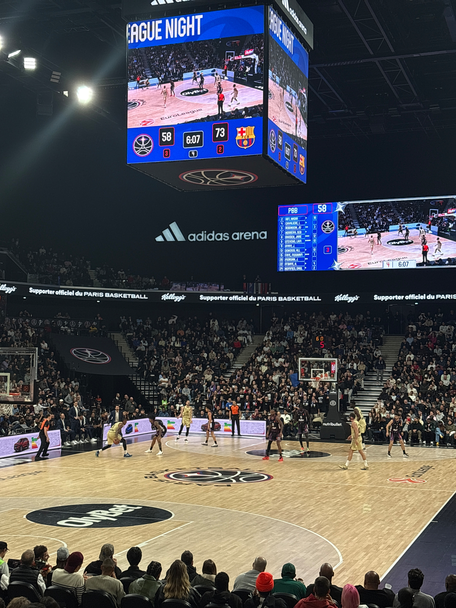 A basketball game is taking place in an indoor arena with spectators watching from the stands and a large screen displaying the score.