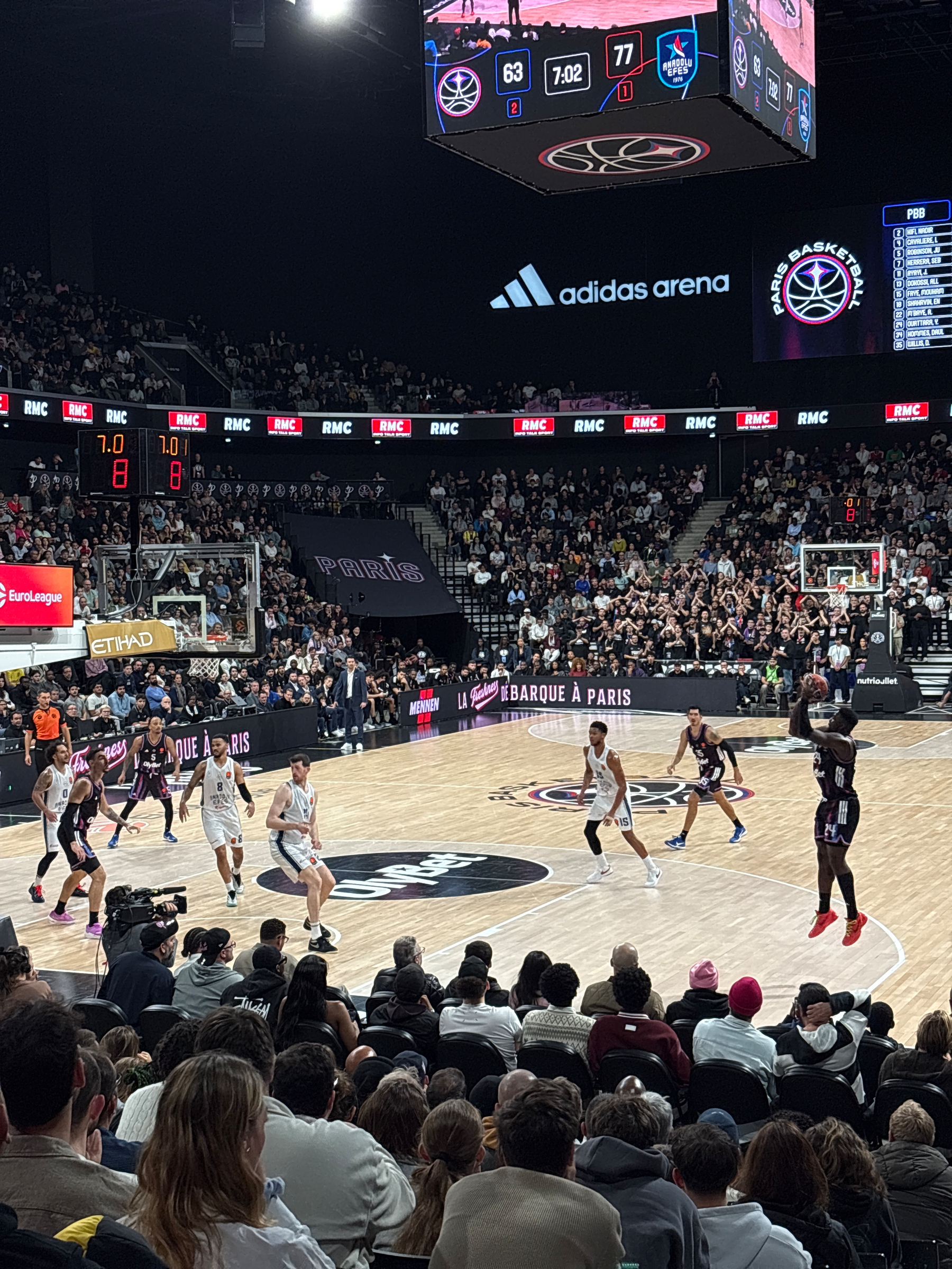 A basketball game is taking place in a packed arena with players in action and a scoreboard showing 63 to 77.