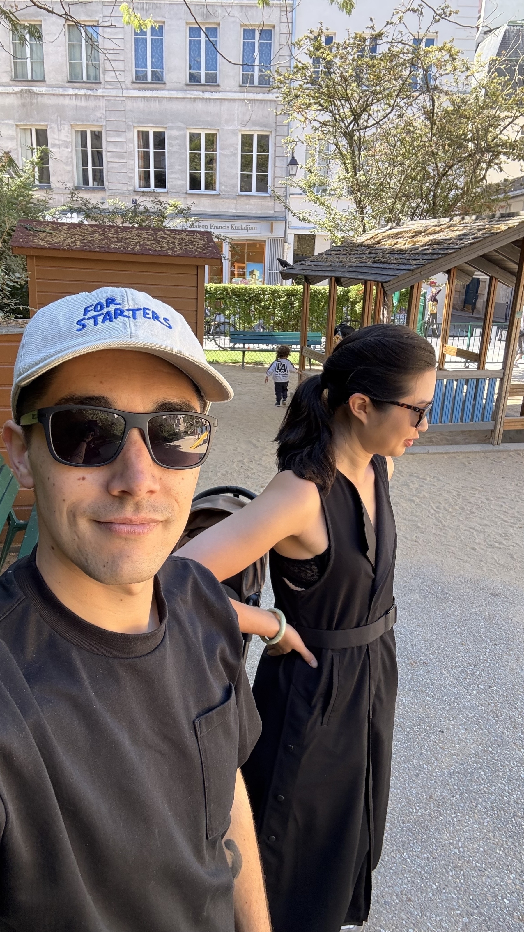 Two people wearing sunglasses are outdoors, with one pushing a stroller near wooden structures.