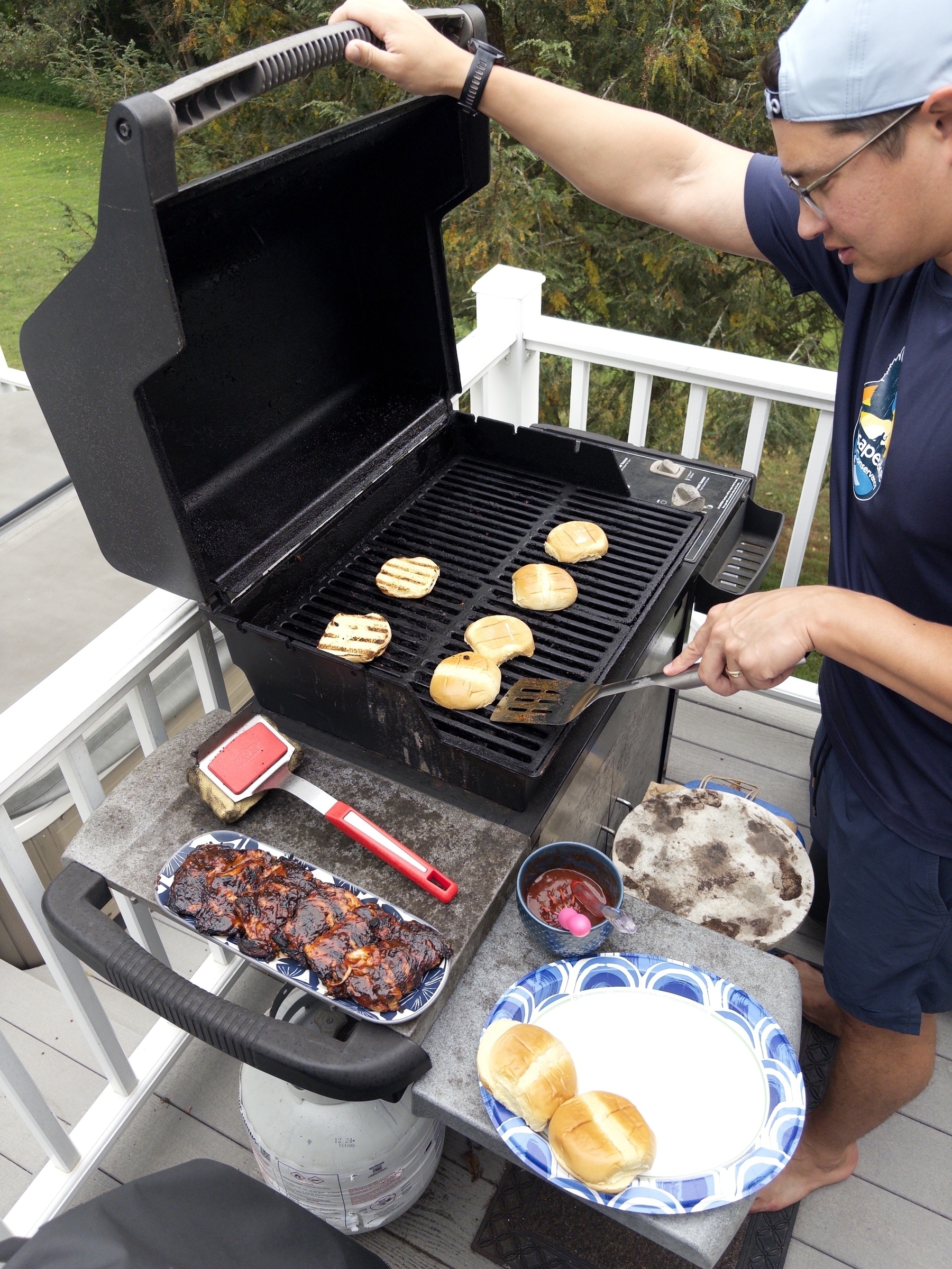 A person is grilling burger buns and chicken on a barbecue grill outside on a deck, with various grilling tools and dishes nearby.
