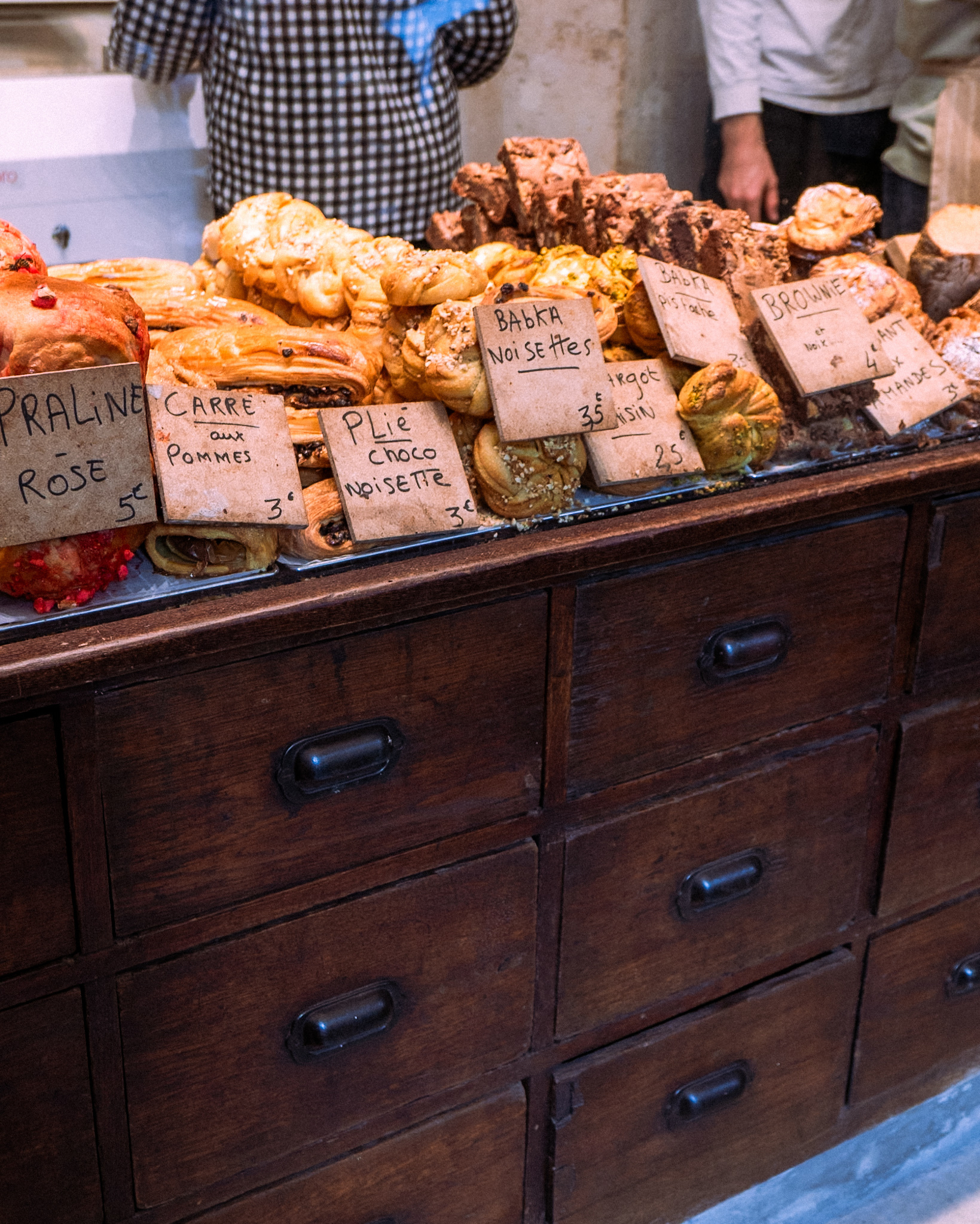 A variety of pastries, including pralines, brownies, and other baked goods, are displayed on an antique wooden counter with handwritten price tags.