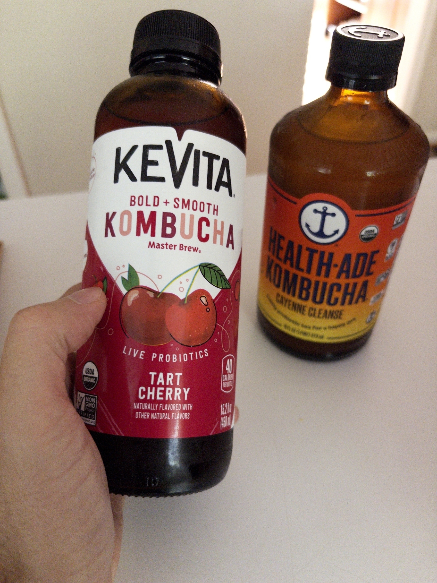 A person is holding a bottle of Kevita Tart Cherry kombucha with another Health-Ade Cayenne Cleanse kombucha bottle in the background.