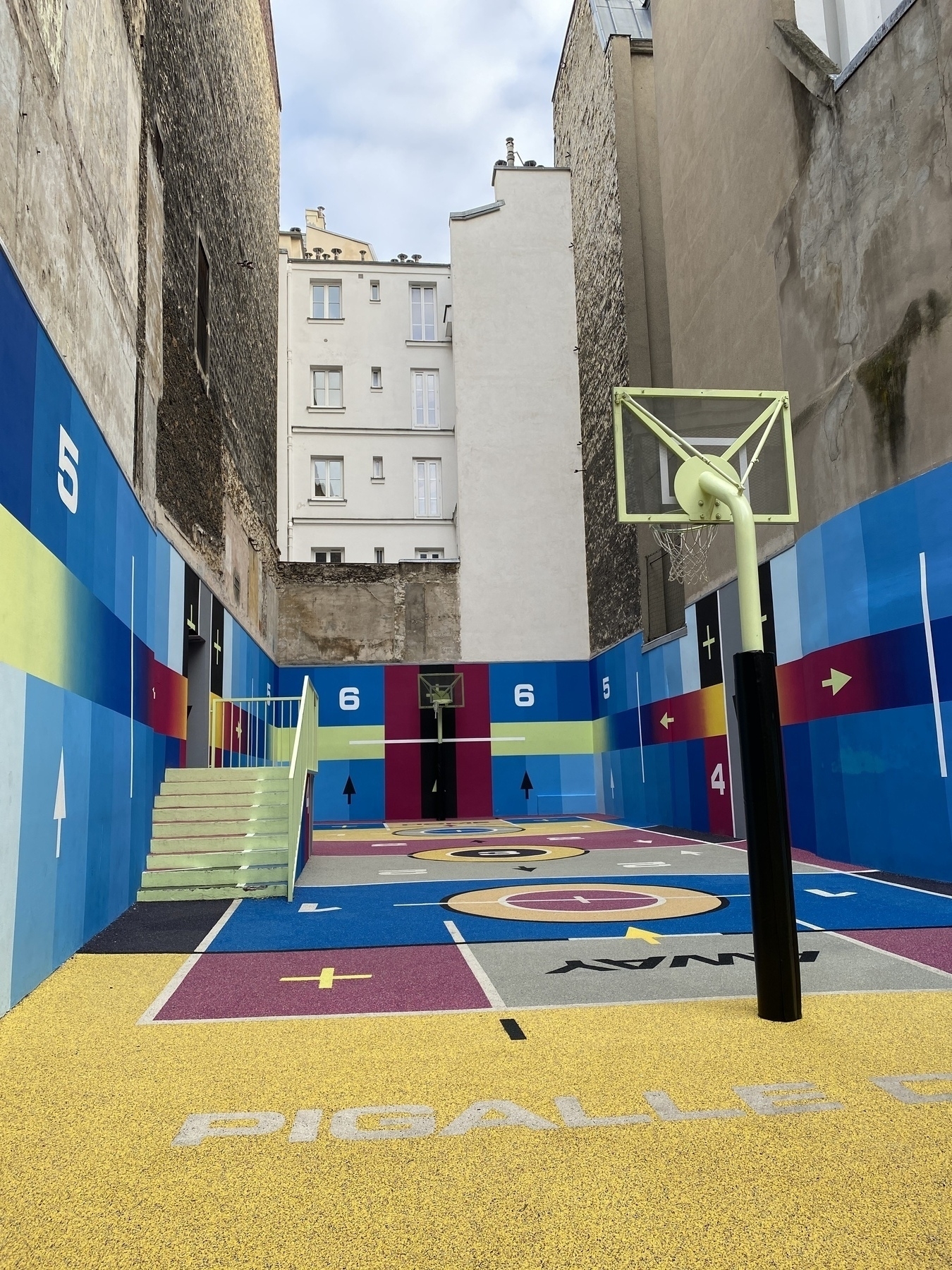 A colorful urban basketball court is surrounded by tall buildings, featuring vibrant geometric designs and pastel hues.