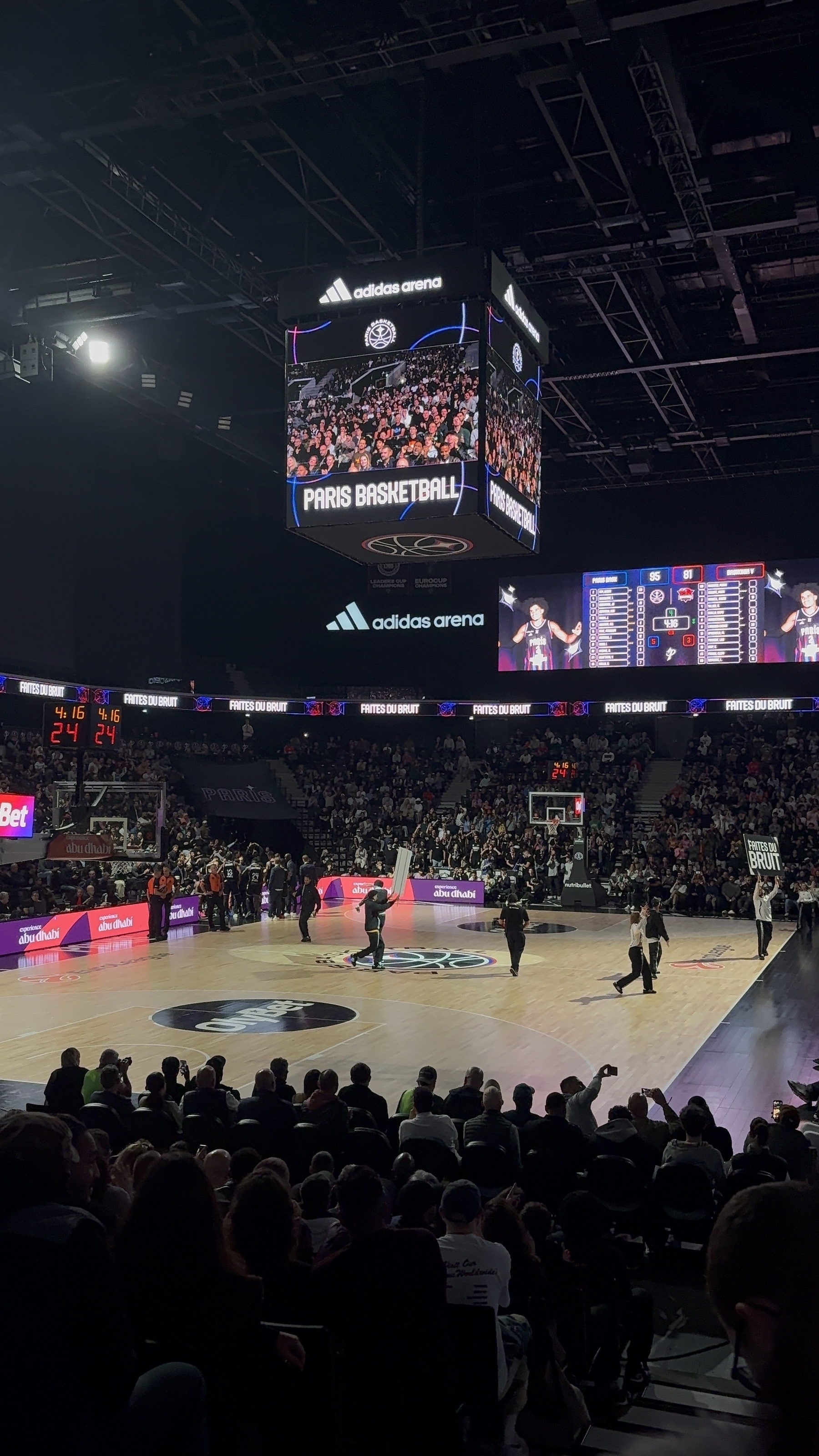 A crowded arena hosts a basketball game, with a scoreboard showing the score and a vibrant display above the court.