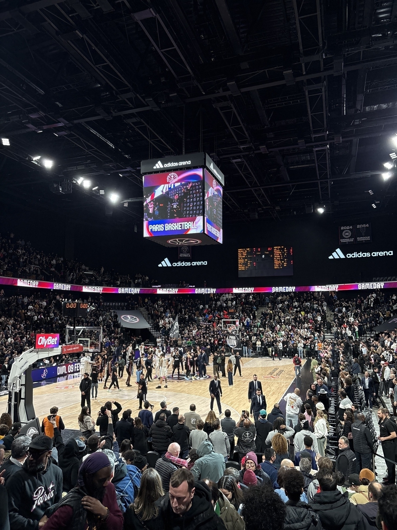 A crowded indoor arena features a basketball game in progress with spectators filling the stands.