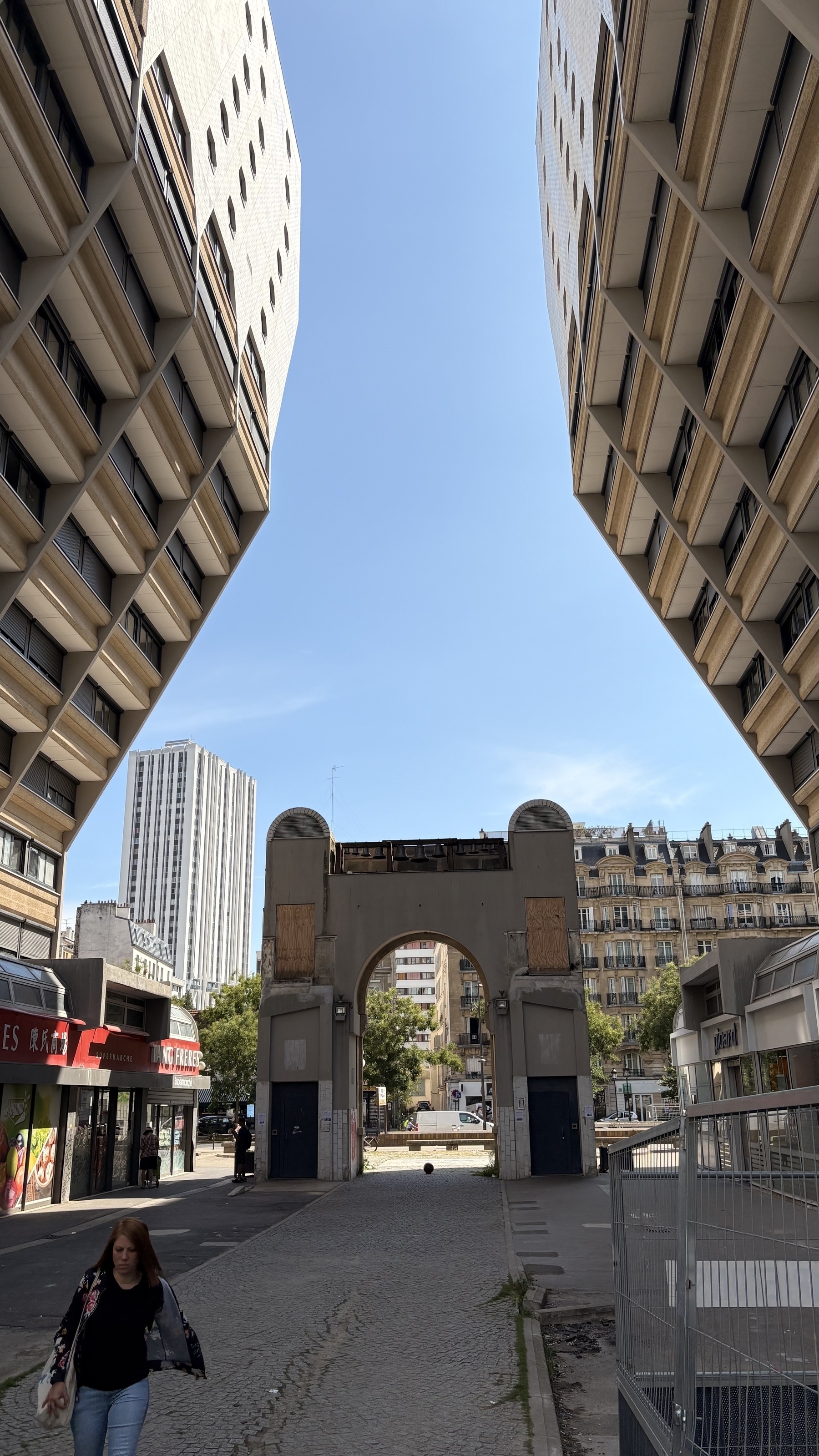 A narrow street with modern, angular buildings on both sides and an arched structure at the end under a clear blue sky.