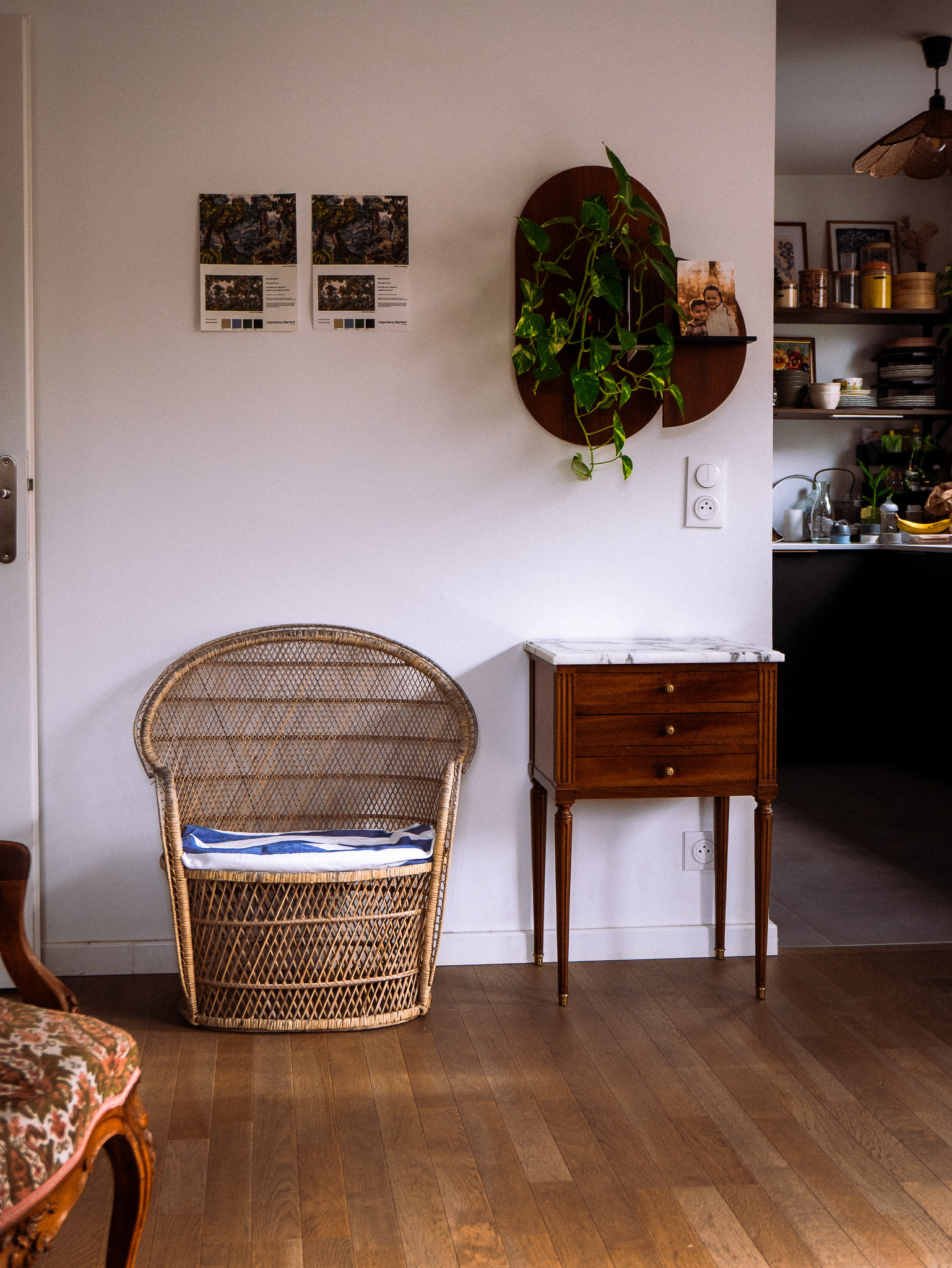 A cozy corner features a wicker chair, a small wooden cabinet with a marble top, and wall-mounted decor with a hanging plant, against a backdrop of a white wall and adjacent kitchen space.
