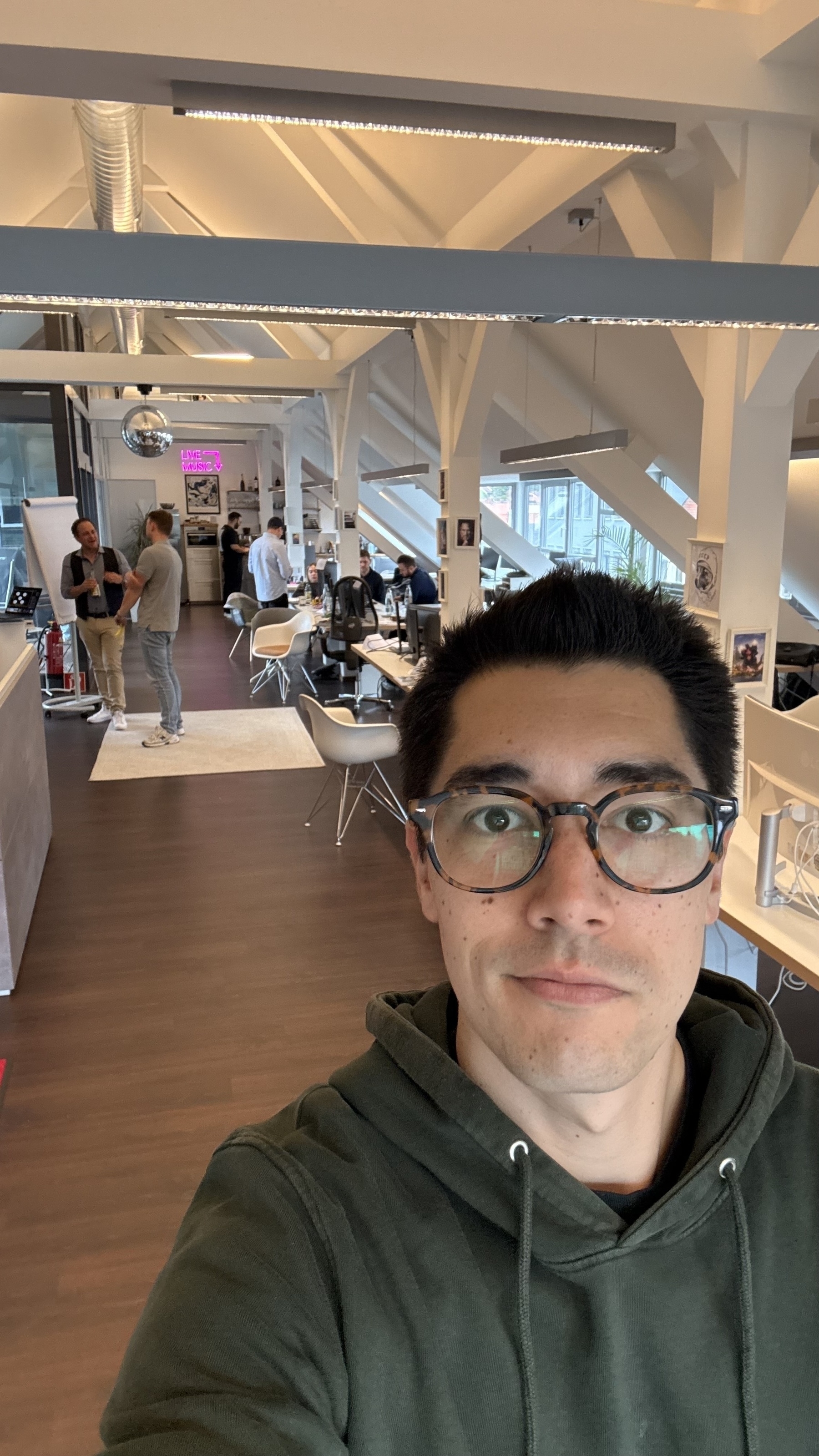 A man in glasses and a green hoodie takes a selfie in an open office with several people working in the background.
