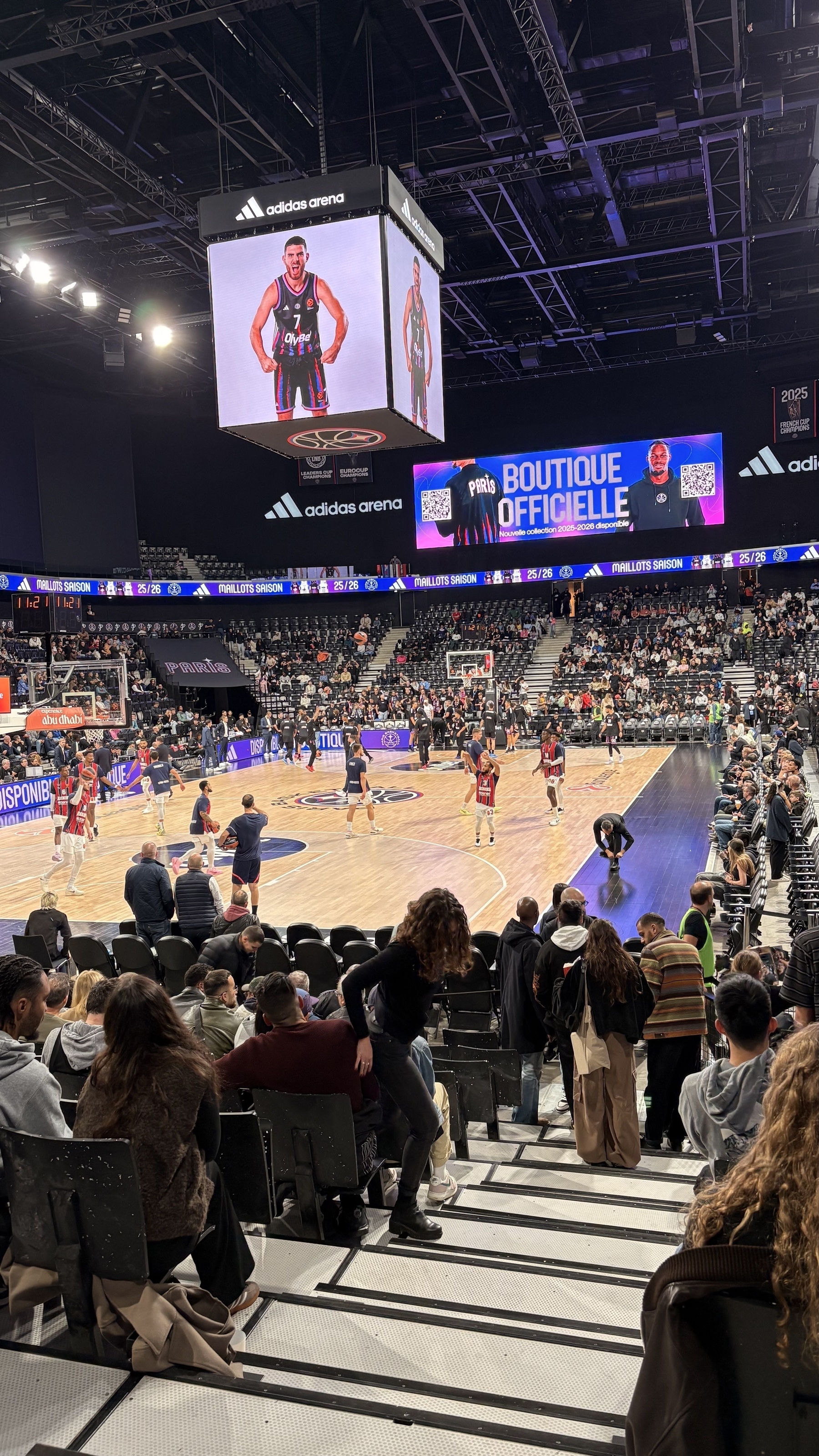 A basketball game is taking place in a large arena with spectators watching from the stands.