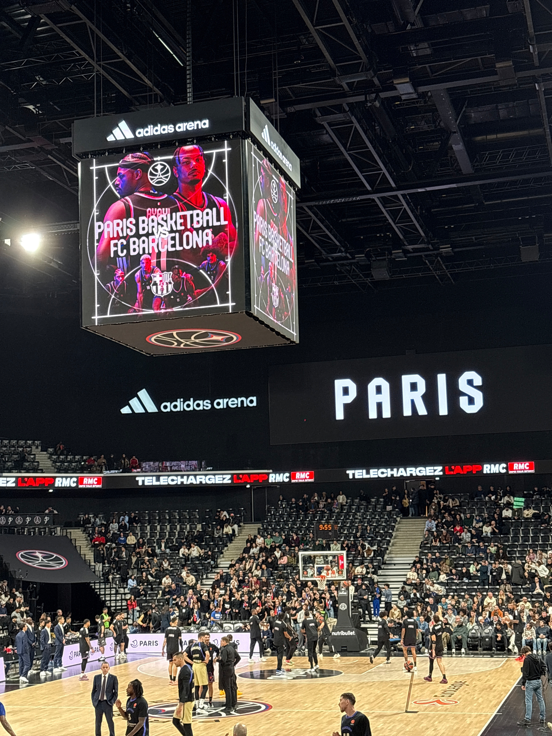 Players warm up on a basketball court under a large digital screen displaying an upcoming match between Paris Basketball and FC Barcelona in an arena.