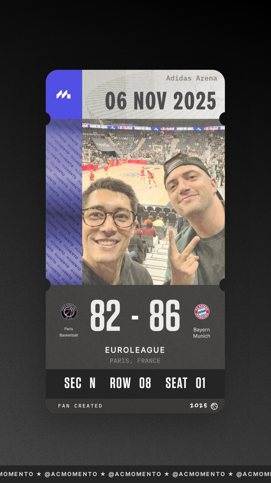 Two people are taking a selfie at a basketball game with the court and players visible in the background.
