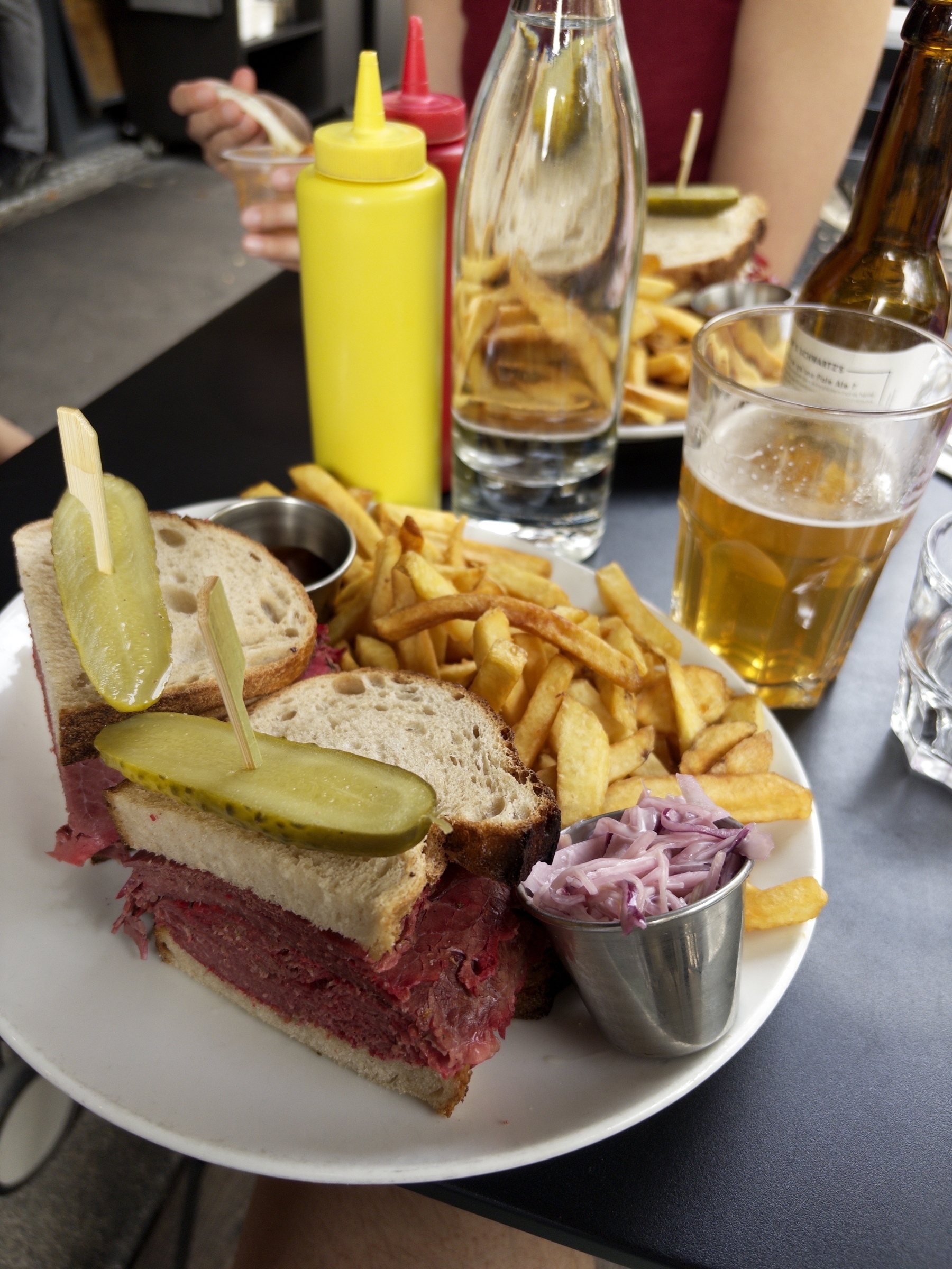 A plate of food featuring a sandwich with pickles, fries, and coleslaw is accompanied by condiments, a glass of beer, and a water bottle on a table.