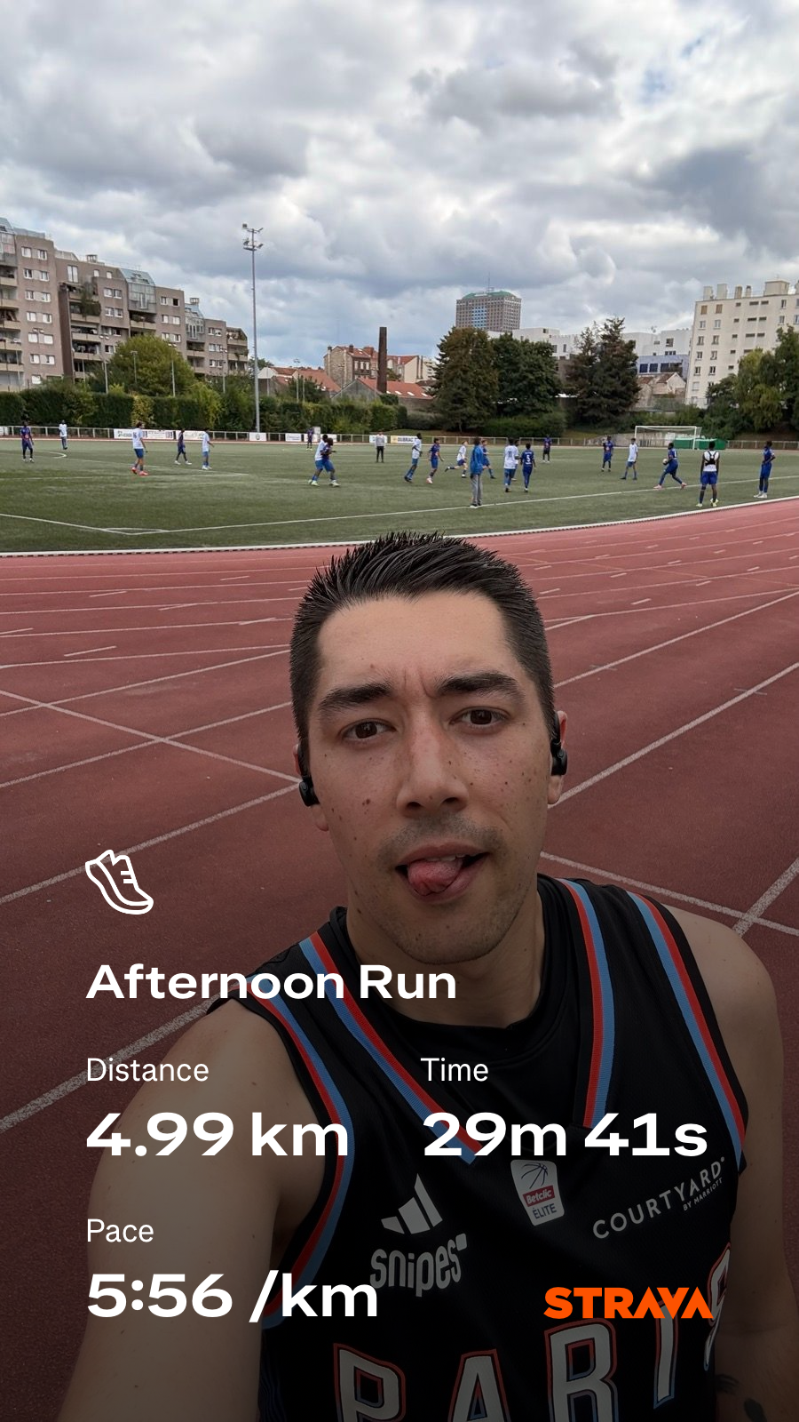 A person is taking a selfie on a field after completing an afternoon run of 4.99 km in 29 minutes and 41 seconds, with a pace of 5:56 per km, while a group of people are seen in the background.
