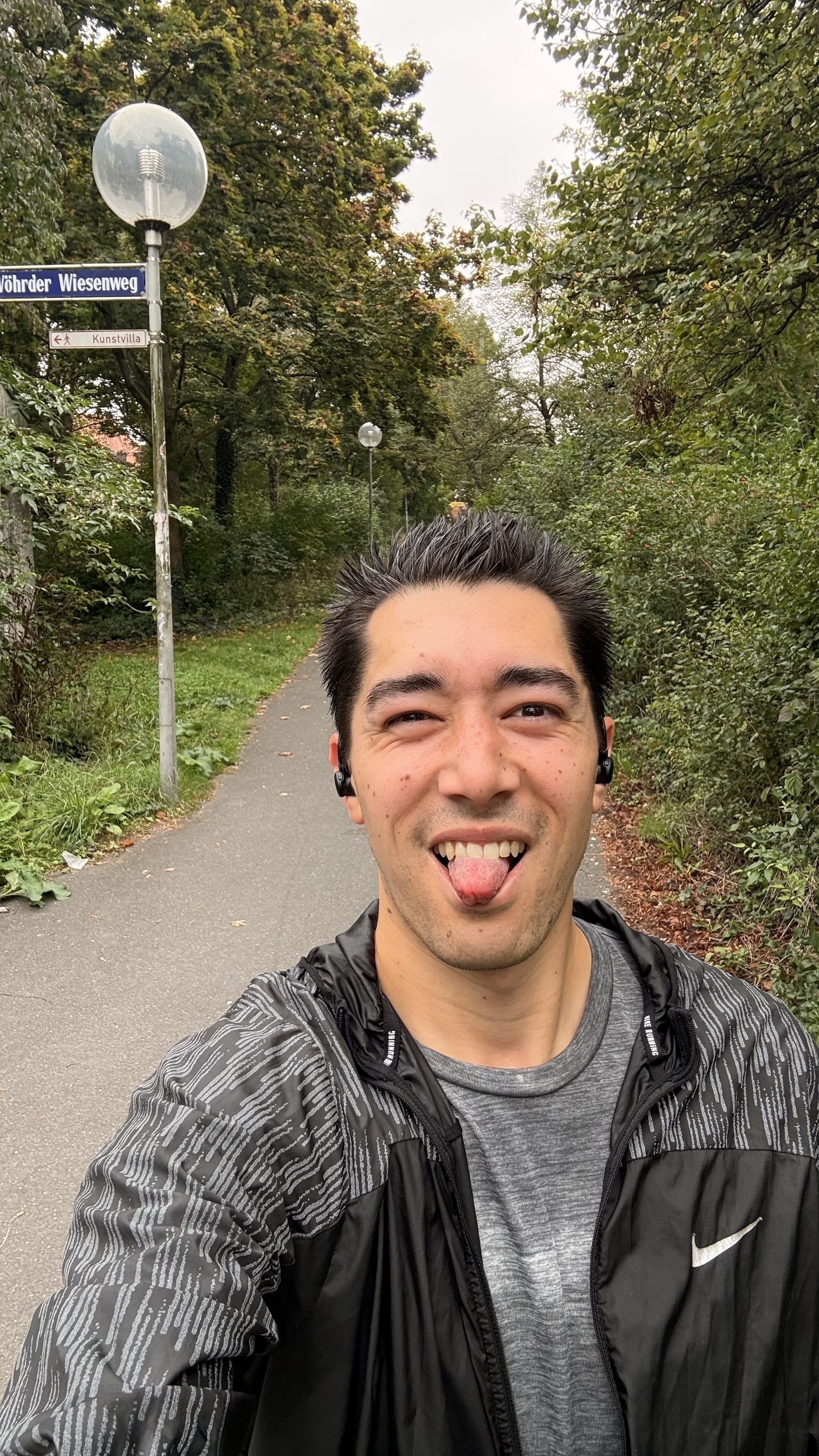 A person wearing headphones and a Nike jacket is taking a selfie on a leafy path with a street lamp and sign in the background.