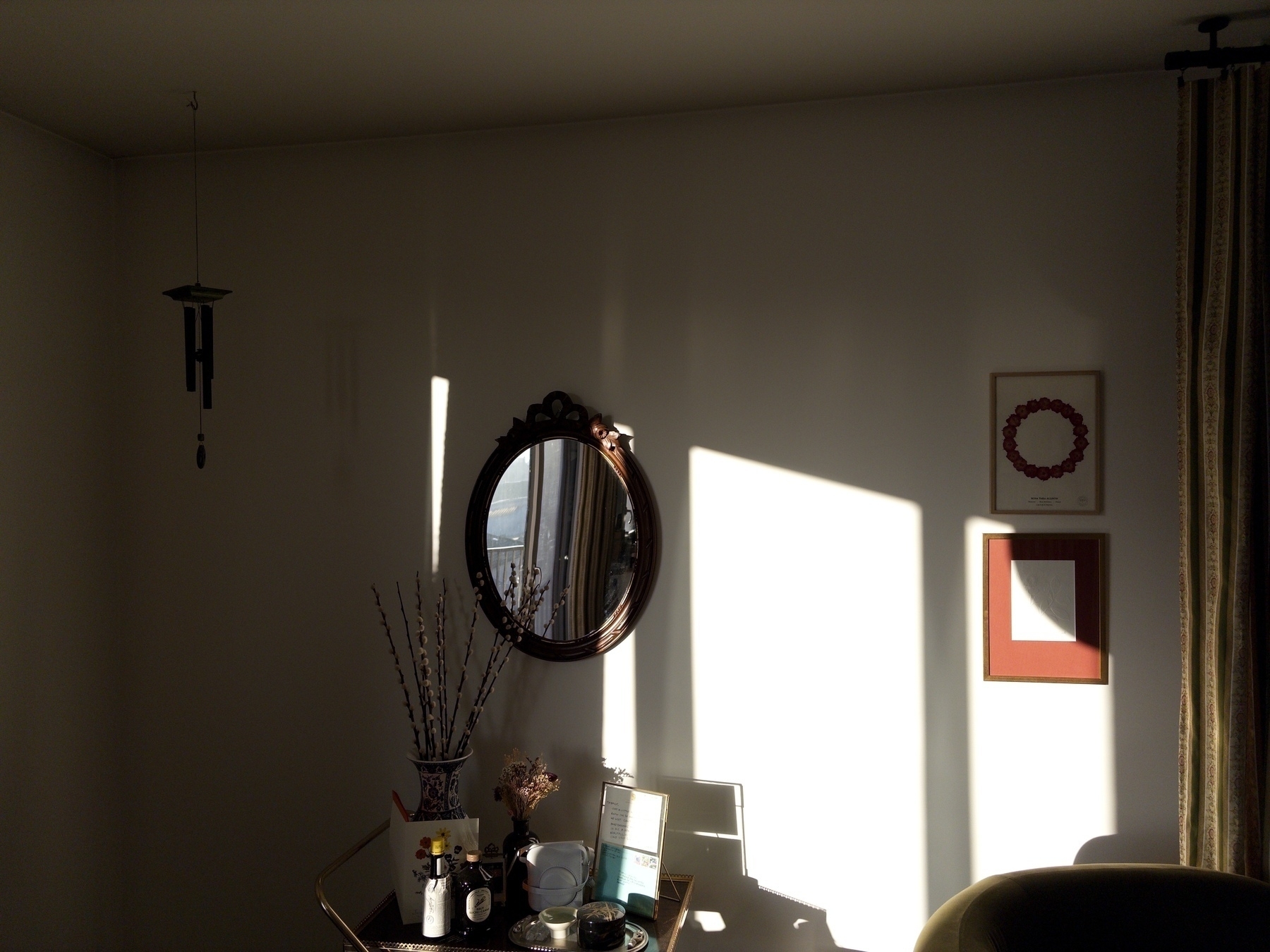 A cozy room corner features a round mirror, a small table with various items, dried plants, and two framed art pieces, all lit by warm sunlight casting shadows on the wall.