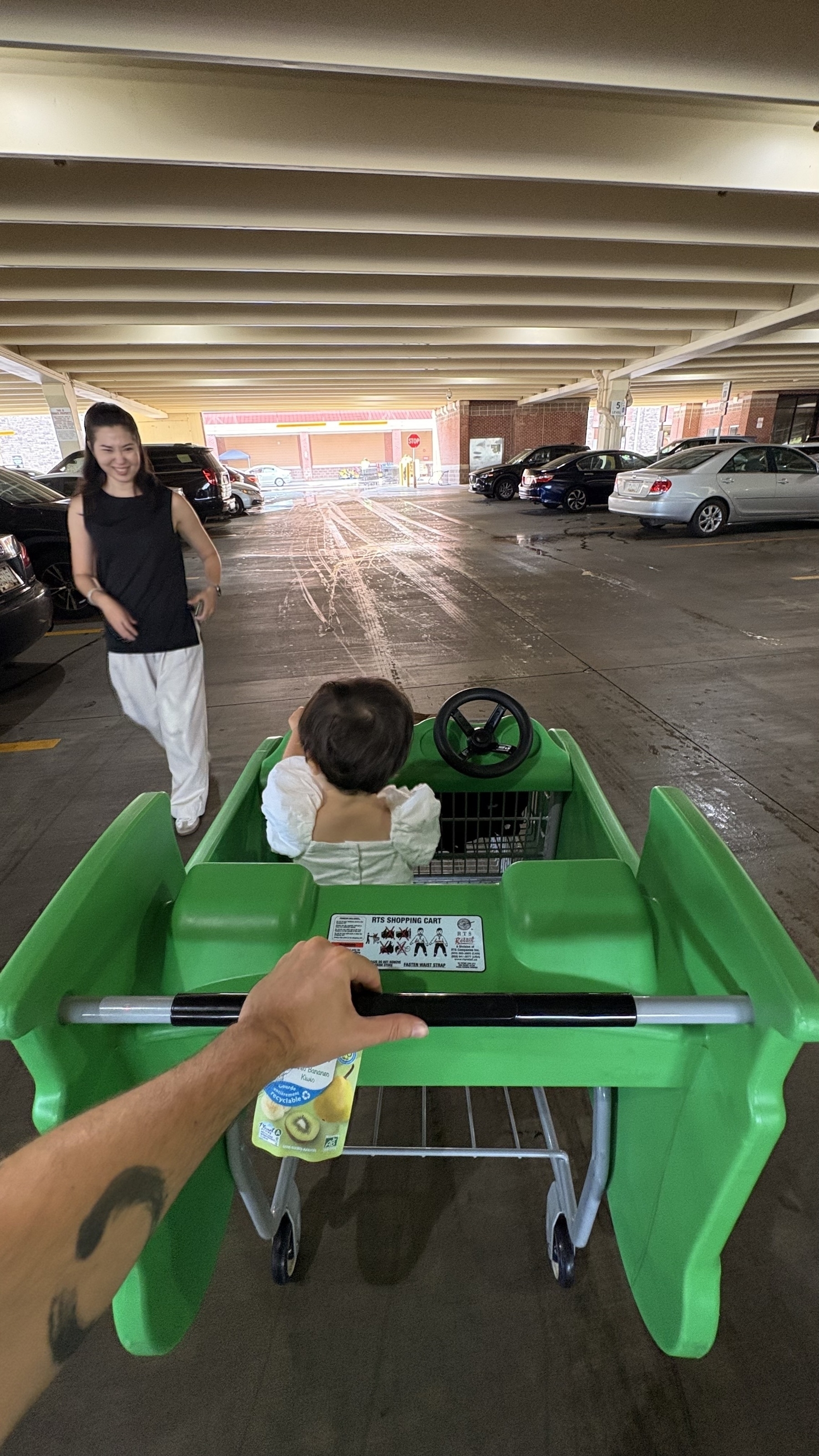 A person is pushing a child in a green shopping cart designed like a car, while another person walks beside them in a parking garage.