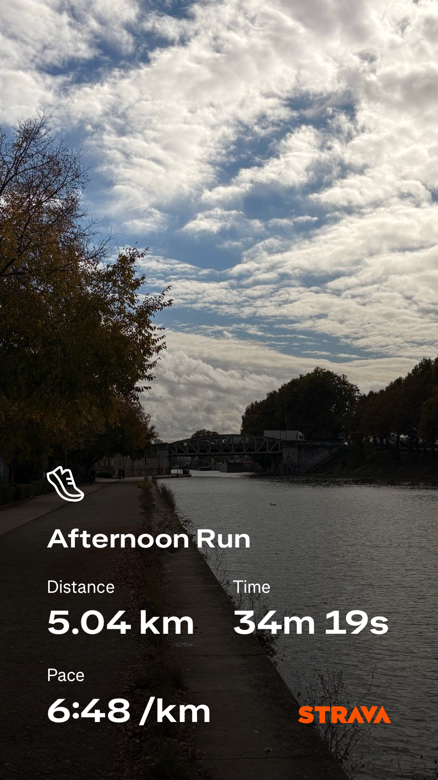 A tranquil riverside path lined with autumn trees stretches under a mostly cloudy sky, displaying the details of an afternoon run.