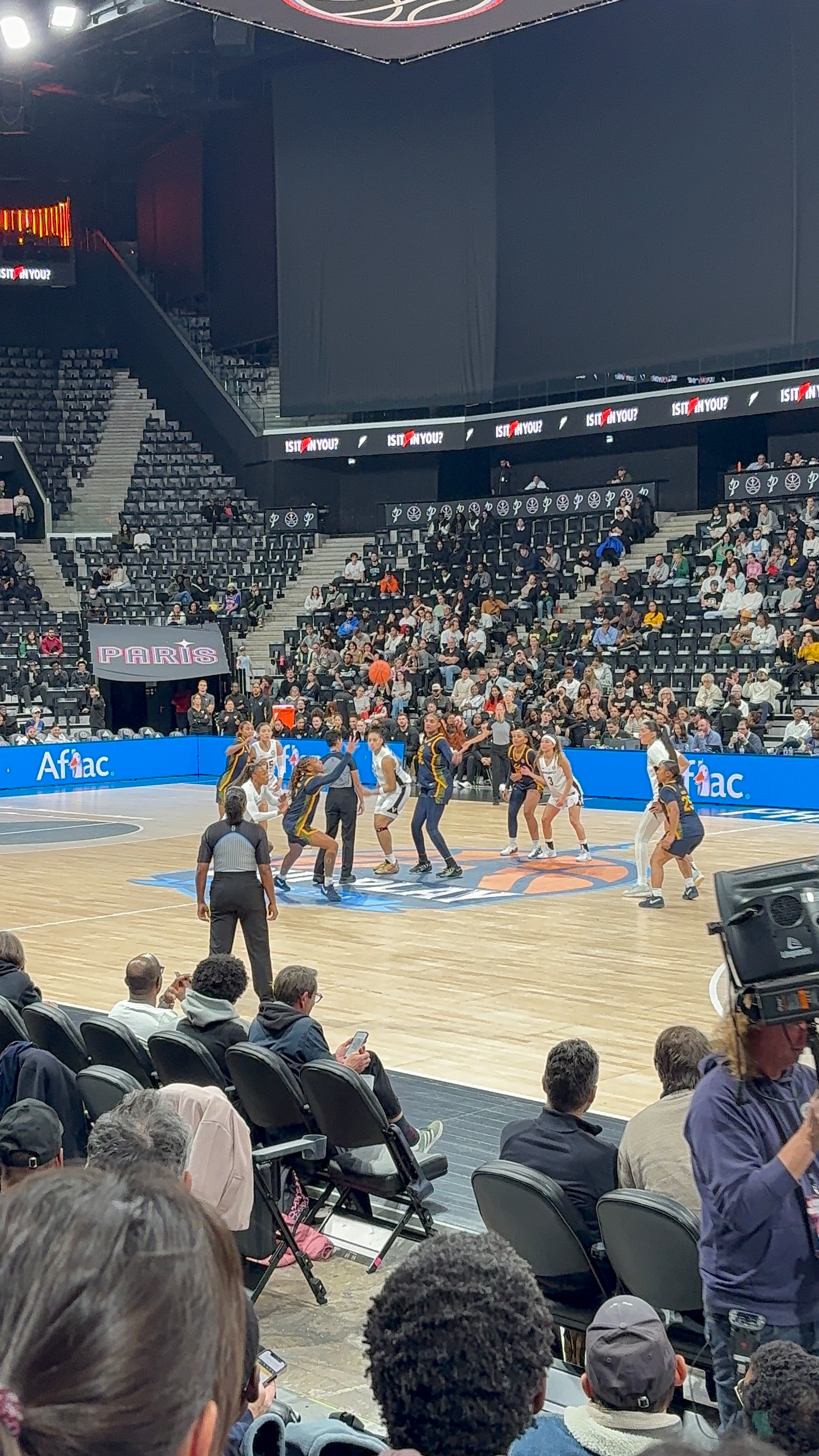 A basketball game is taking place on a court in an indoor arena with players ready for a tip-off and spectators watching.