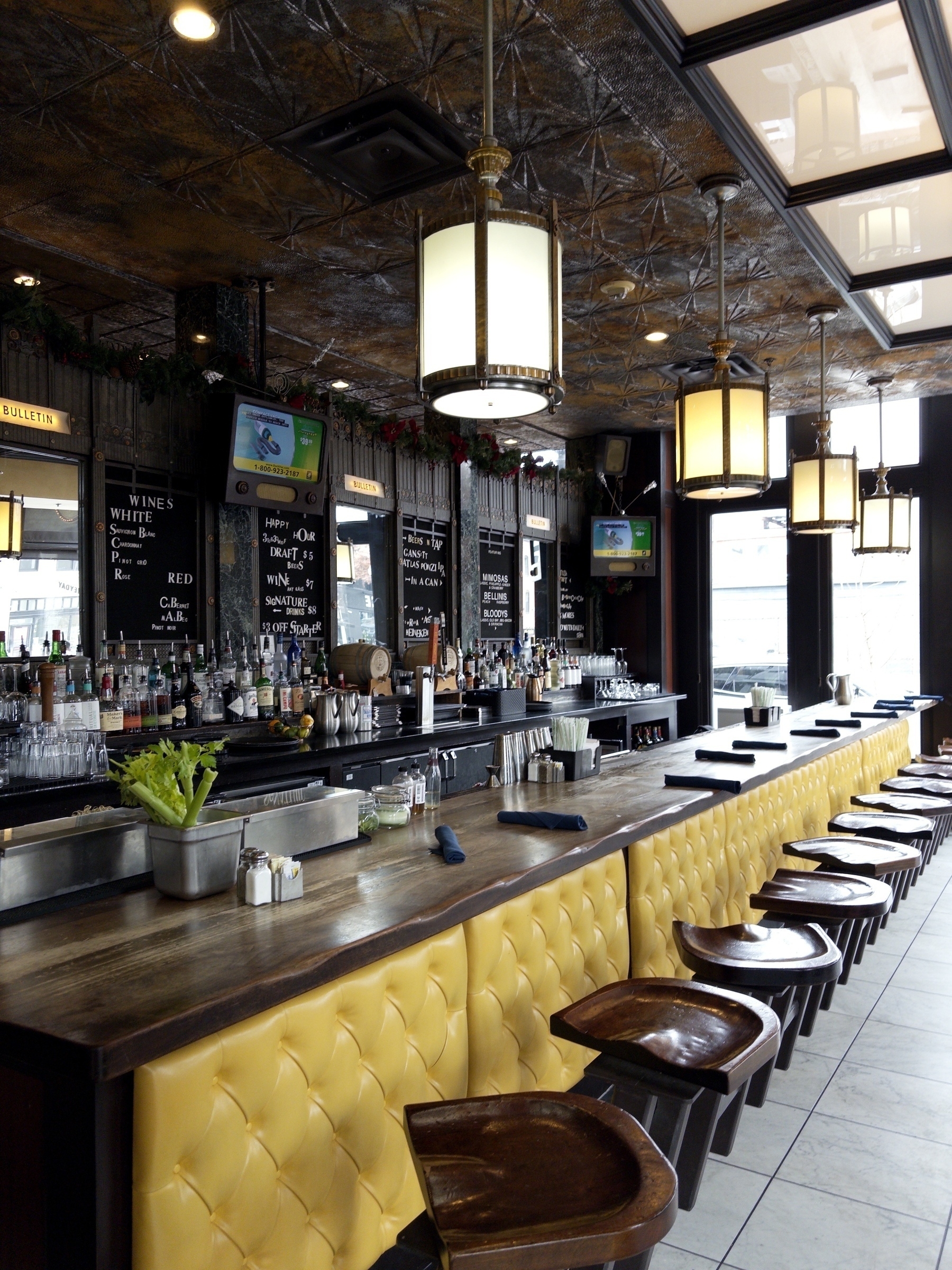 A trendy bar features a long counter with yellow tufted seating, stylish hanging lights, and a variety of drinks displayed on shelves behind it.